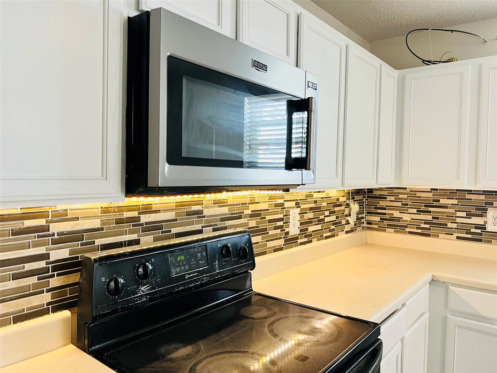 White shaker-style cabinetry, mosaic tile backsplash, under-cabinet lighting, stainless steel over-the-range microwave, and solid surface countertops