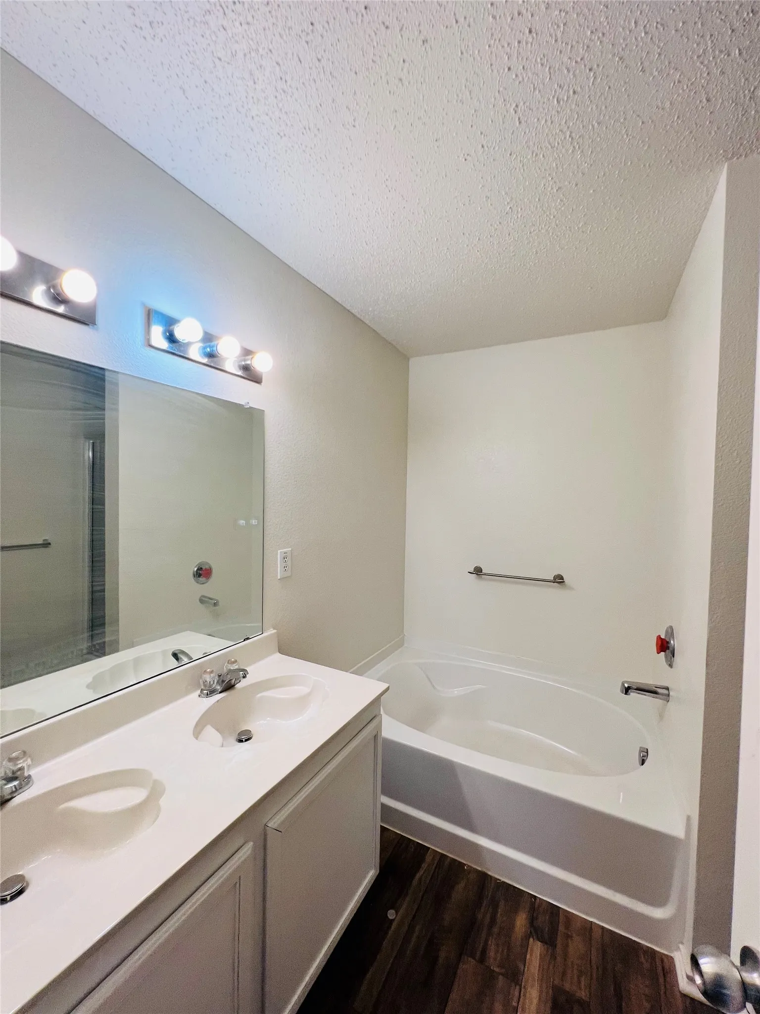 Dual vanity with integrated sinks, wood-finish flooring, built-in bathtub, and wall-mounted lighting fixtures