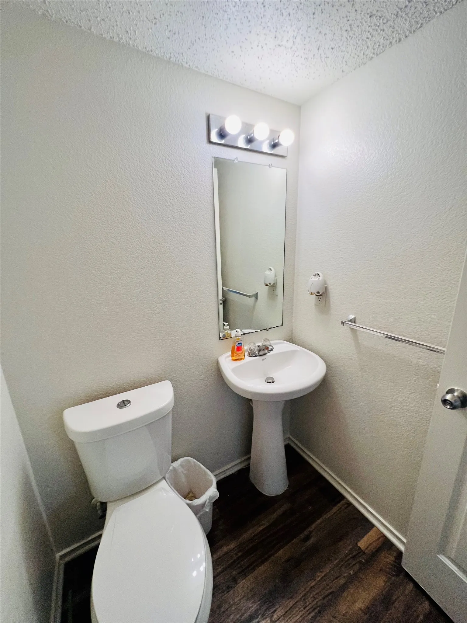 Pedestal sink, wall-mounted mirror, overhead vanity lighting, wood-finish flooring, and a white toilet