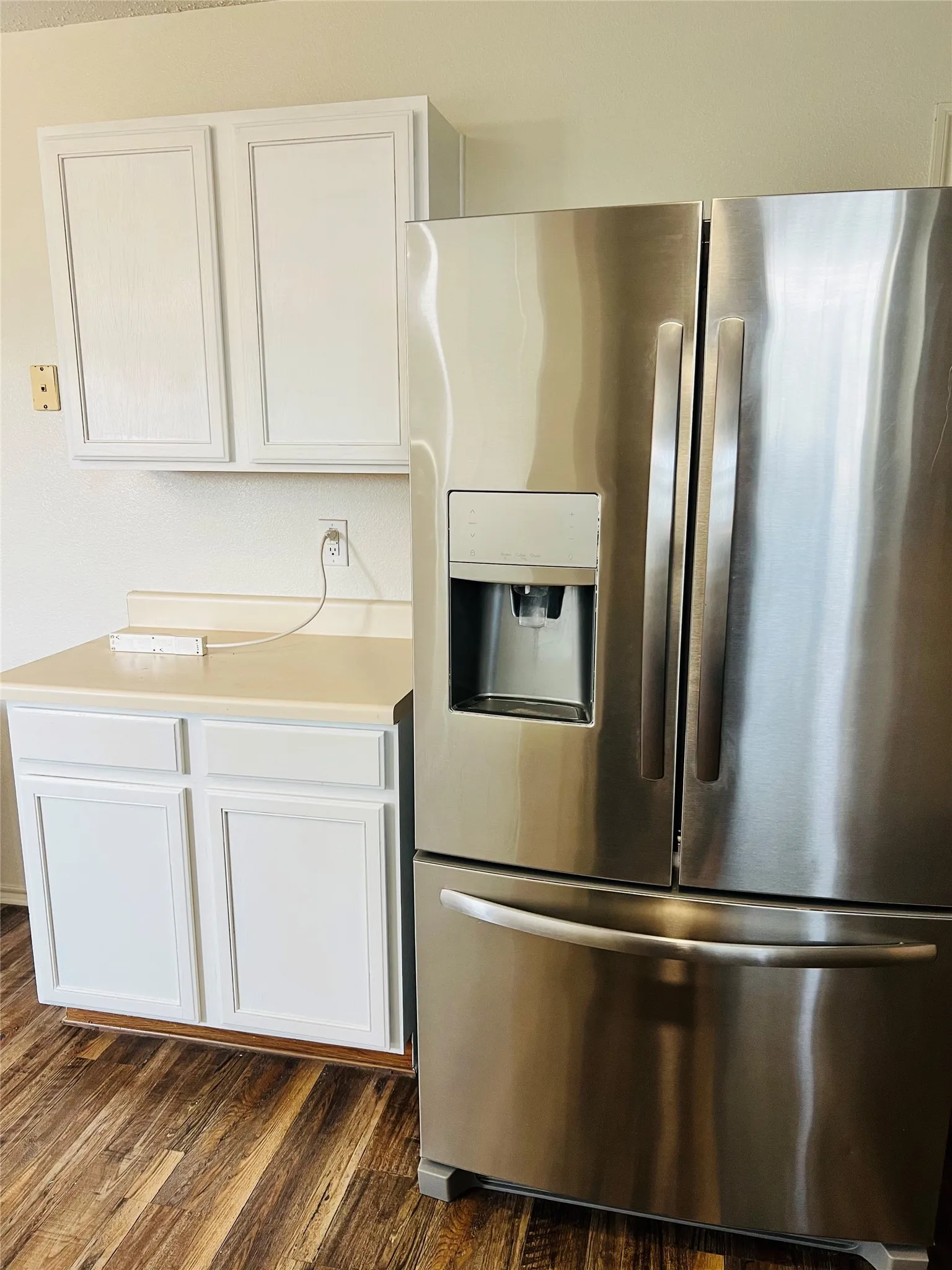 Stainless steel French door refrigerator with an external ice and water dispenser, white cabinetry, light-toned countertop, and wood-finish flooring