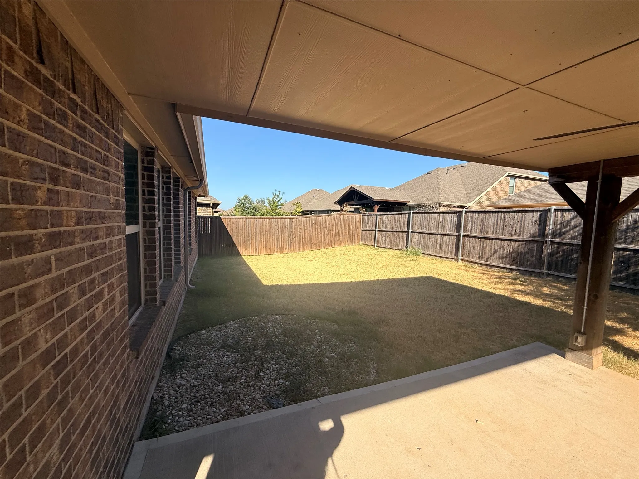 Covered patio with concrete flooring, supported by a wood post structure, overlooking a fenced backyard with a grass lawn