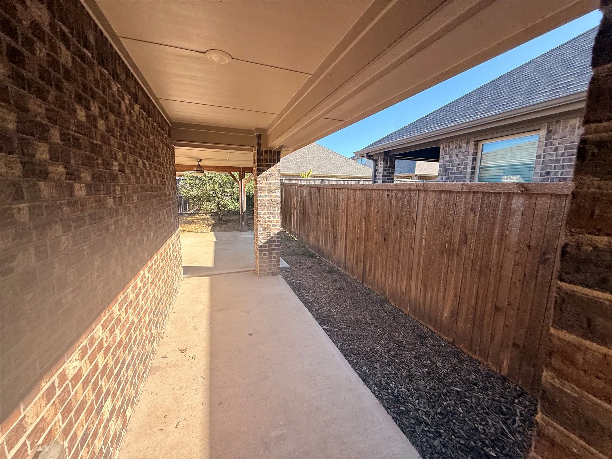 Covered patio with a concrete walkway, brick exterior, and wood fencing