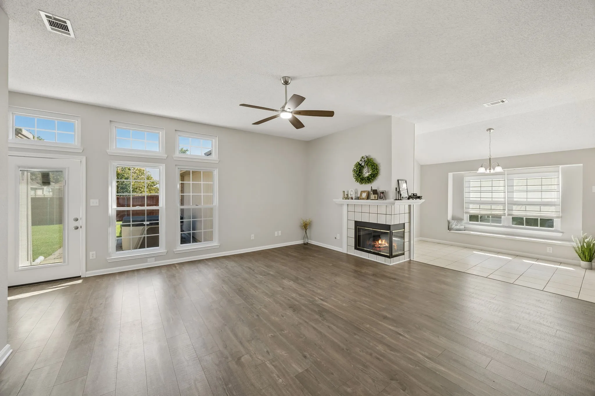 Bright and spacious living room featuring warm wood-look flooring, a corner fireplace, ceiling fan, and a dramatic wall of windows and transoms overlooking the backyard.