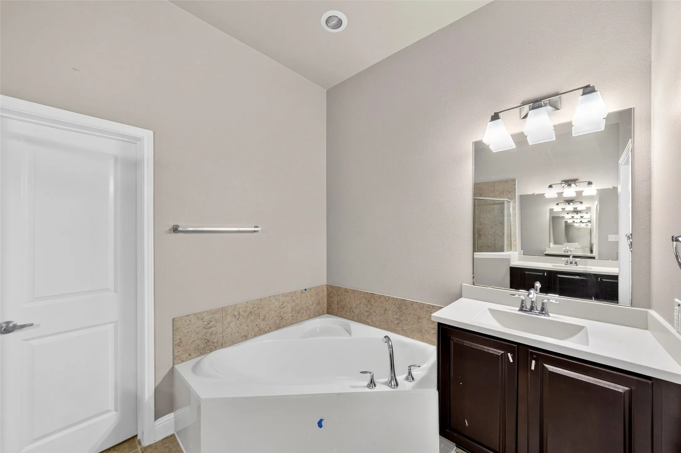 Corner soaking tub with deck-mounted chrome faucet, single vanity with dark wood-finish cabinetry, integrated sink, and chrome fixtures
