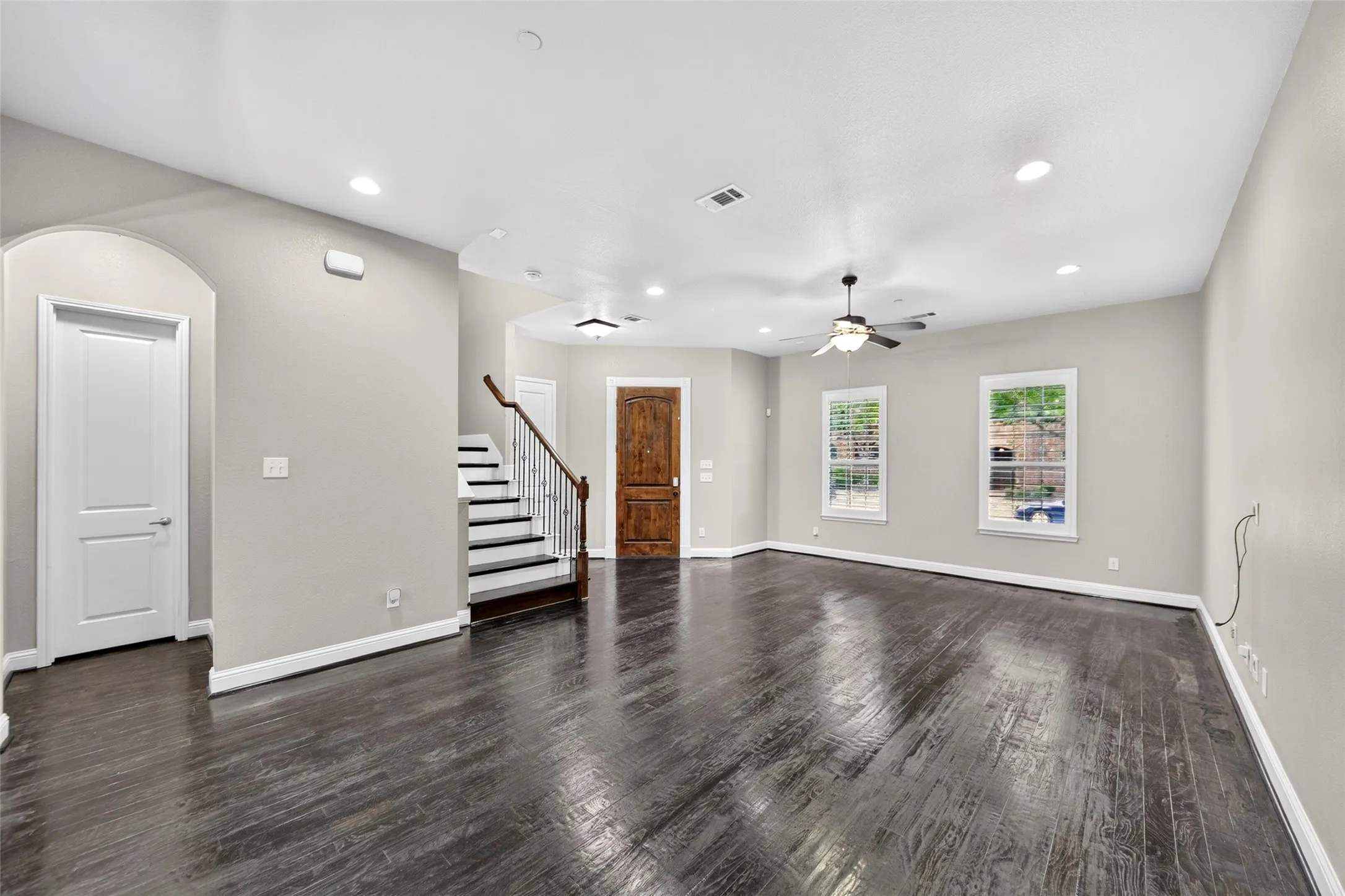 Dark wood-finish flooring, recessed lighting, ceiling fan, natural light from multiple windows, and a staircase with an ornamental railing