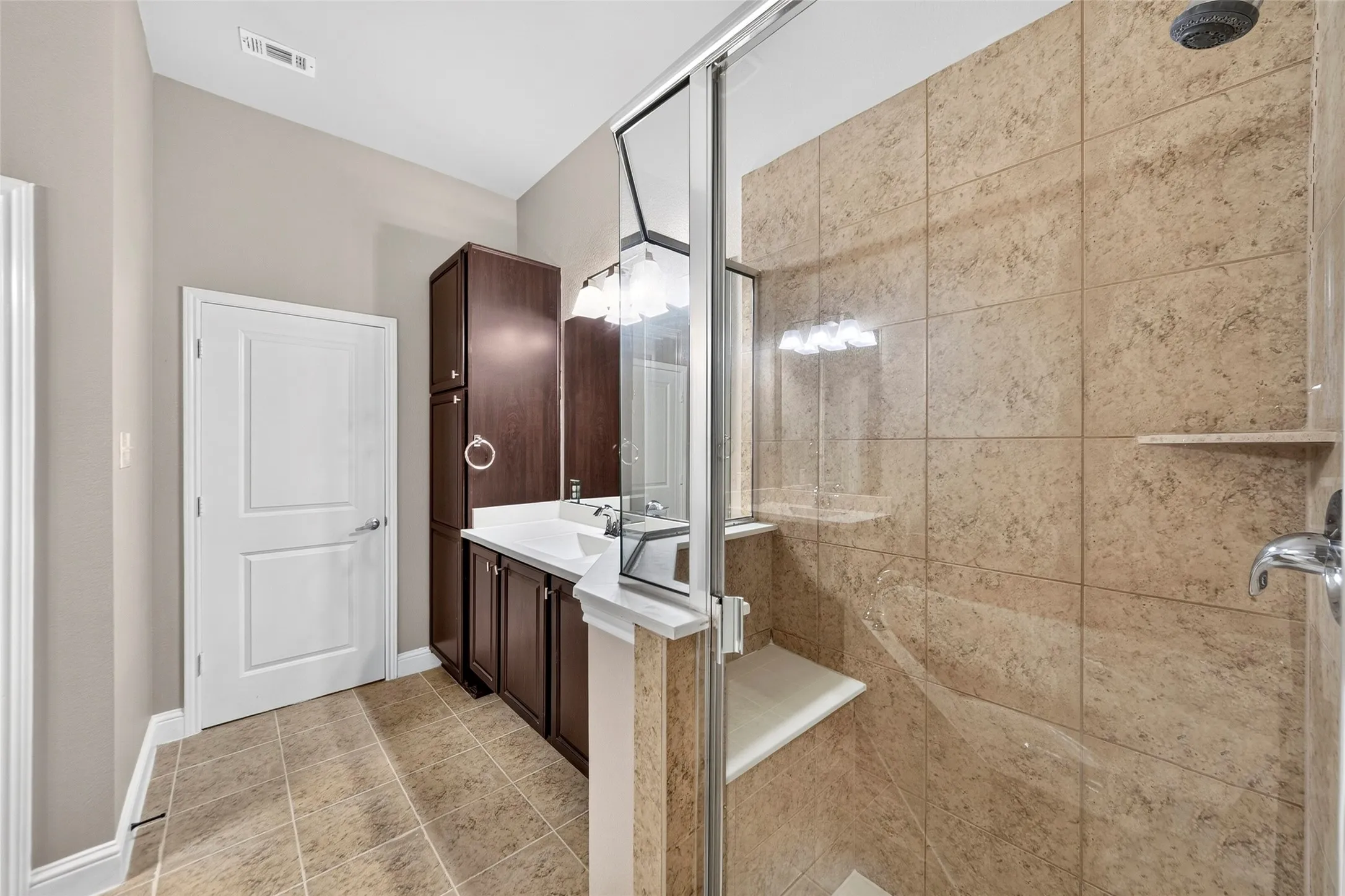 Walk-in shower with tiled walls and an integrated bench, dark wood-finish vanity with white countertop and integrated sink, chrome fixtures, and tiled flooring