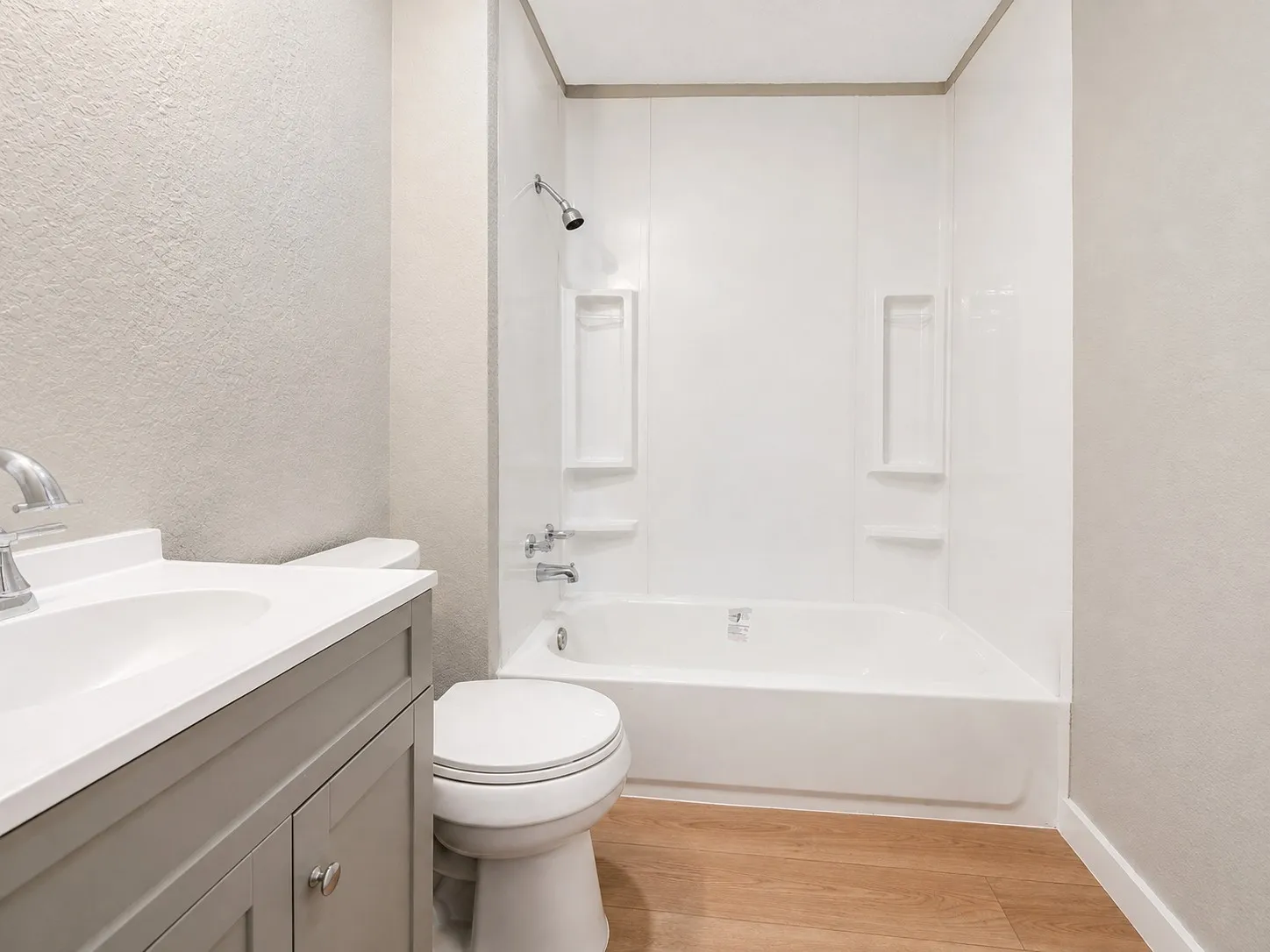 Bathroom featuring a gray vanity with a white countertop, chrome fixtures, a white toilet, a white tub and shower surround, and wood-finish flooring
