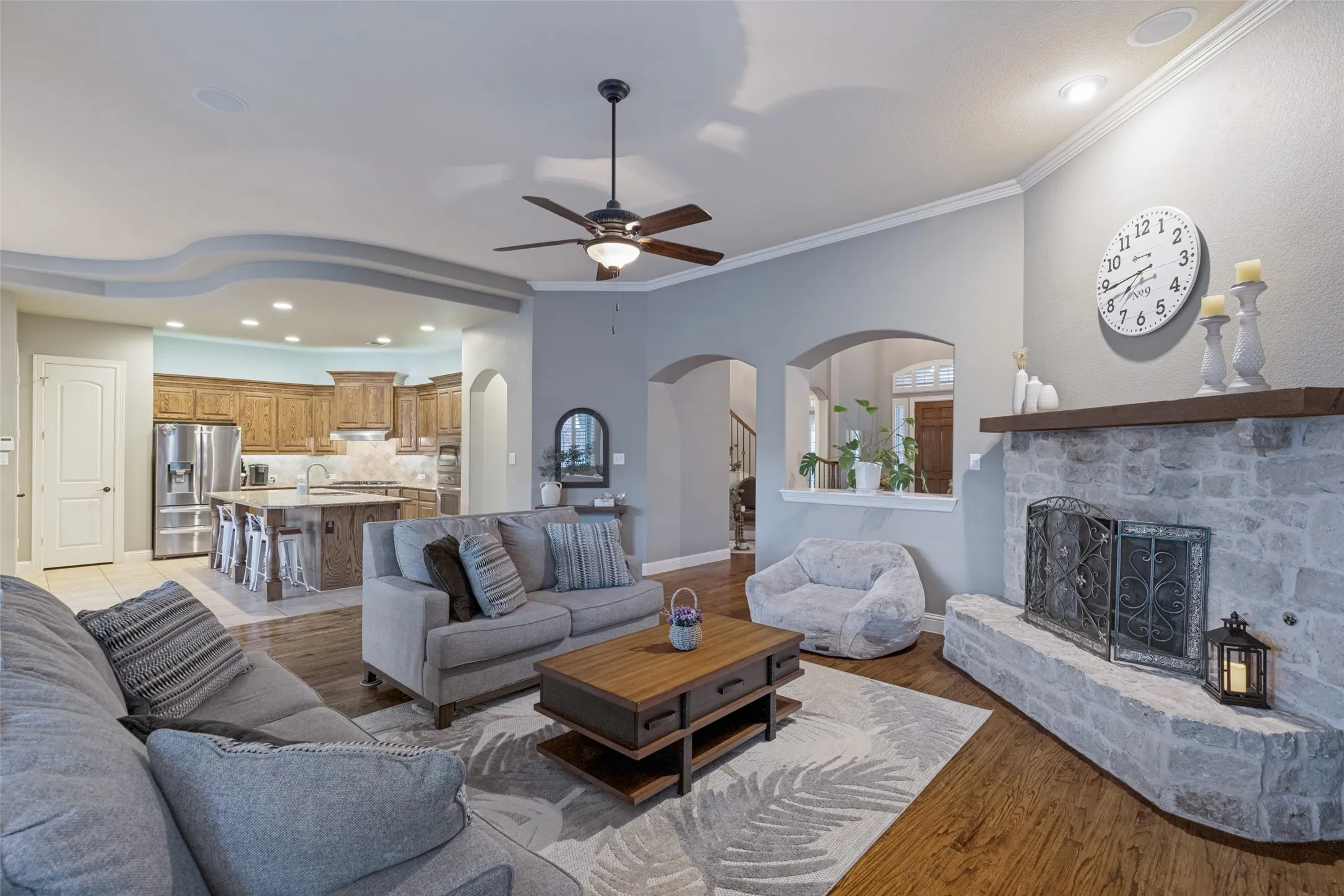 Spacious living area featuring a stone-clad fireplace with a wood mantel, wood-finish flooring, and crown molding
