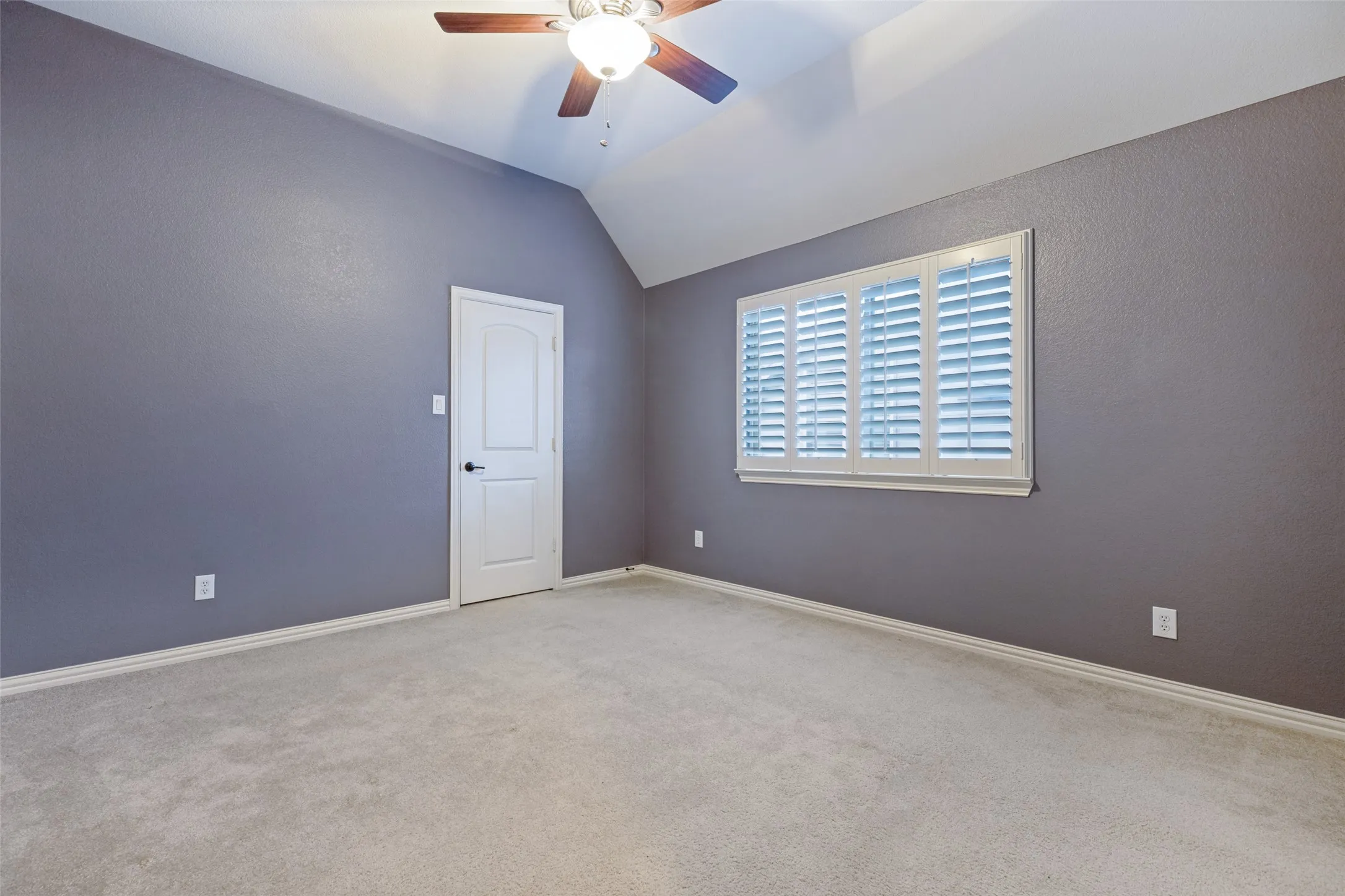 Vaulted ceiling with a ceiling fan, window with plantation shutters, light gray wall finish, and light-colored carpeting