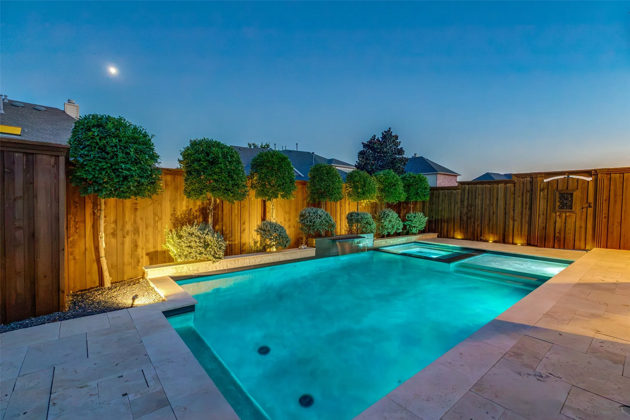 Swimming pool with integrated spa and waterfall feature, surrounded by paver decking and a wood privacy fence