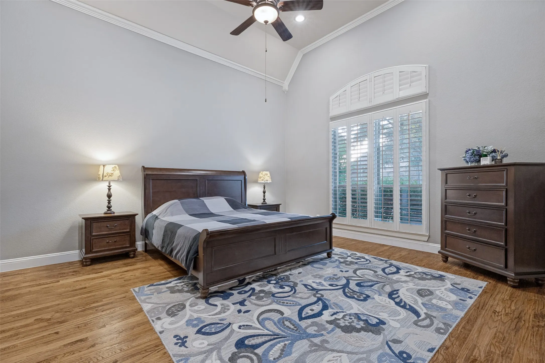 Vaulted ceiling with crown molding, wood-finish flooring, ceiling fan, and arched window with plantation shutters