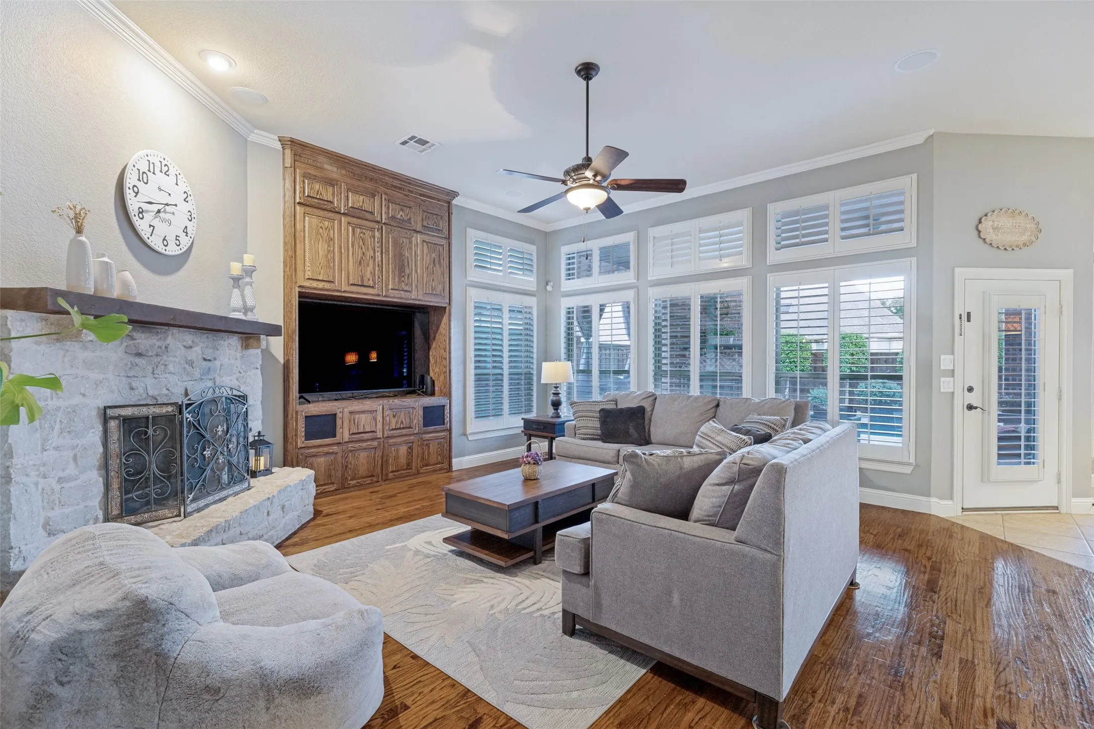 Stone-clad fireplace with mantel, built-in wood cabinetry, wood-finish flooring, ceiling fan, and multiple windows with plantation shutters
