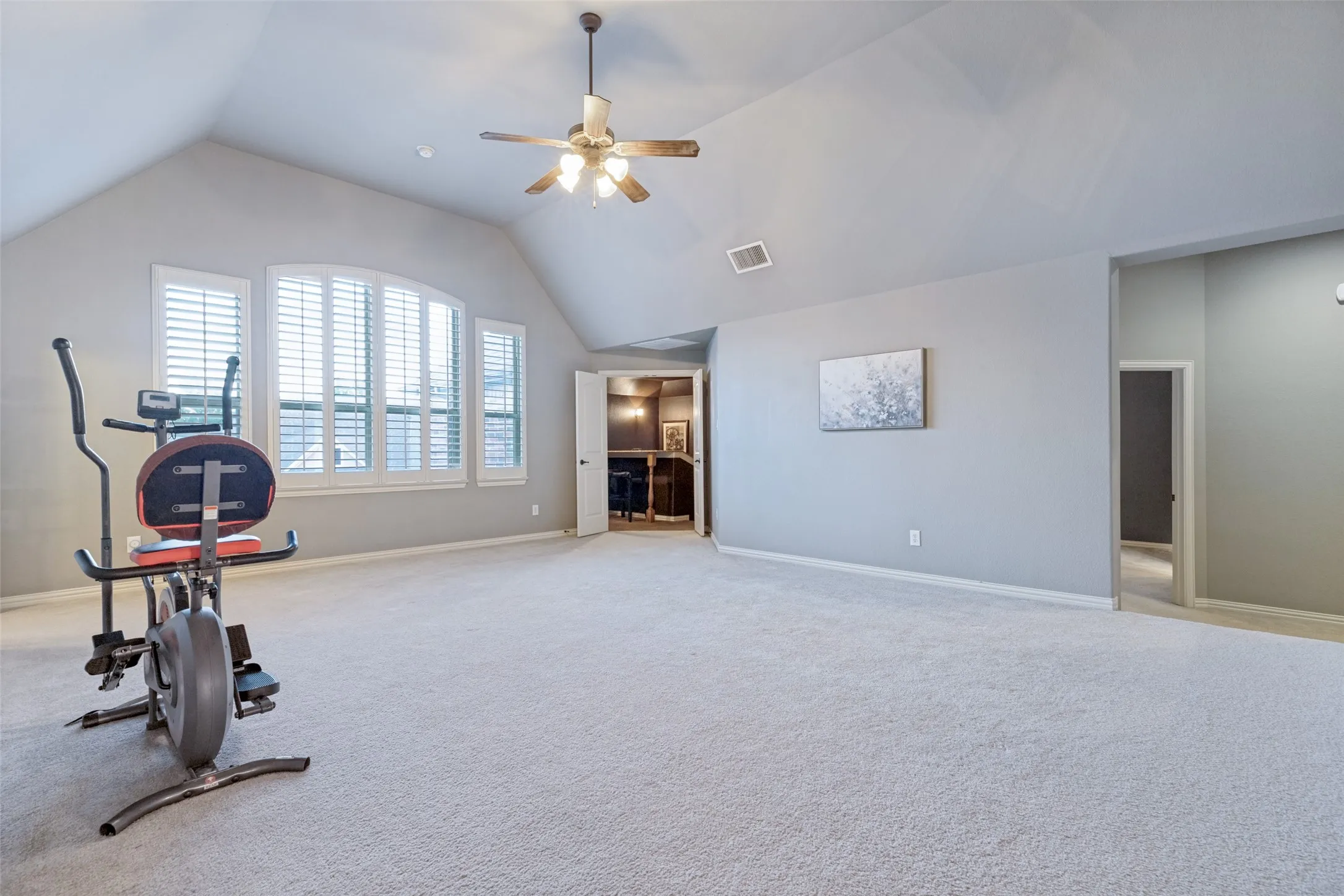 Expansive carpeted room featuring vaulted ceilings, a ceiling fan, and multiple windows with plantation shutters