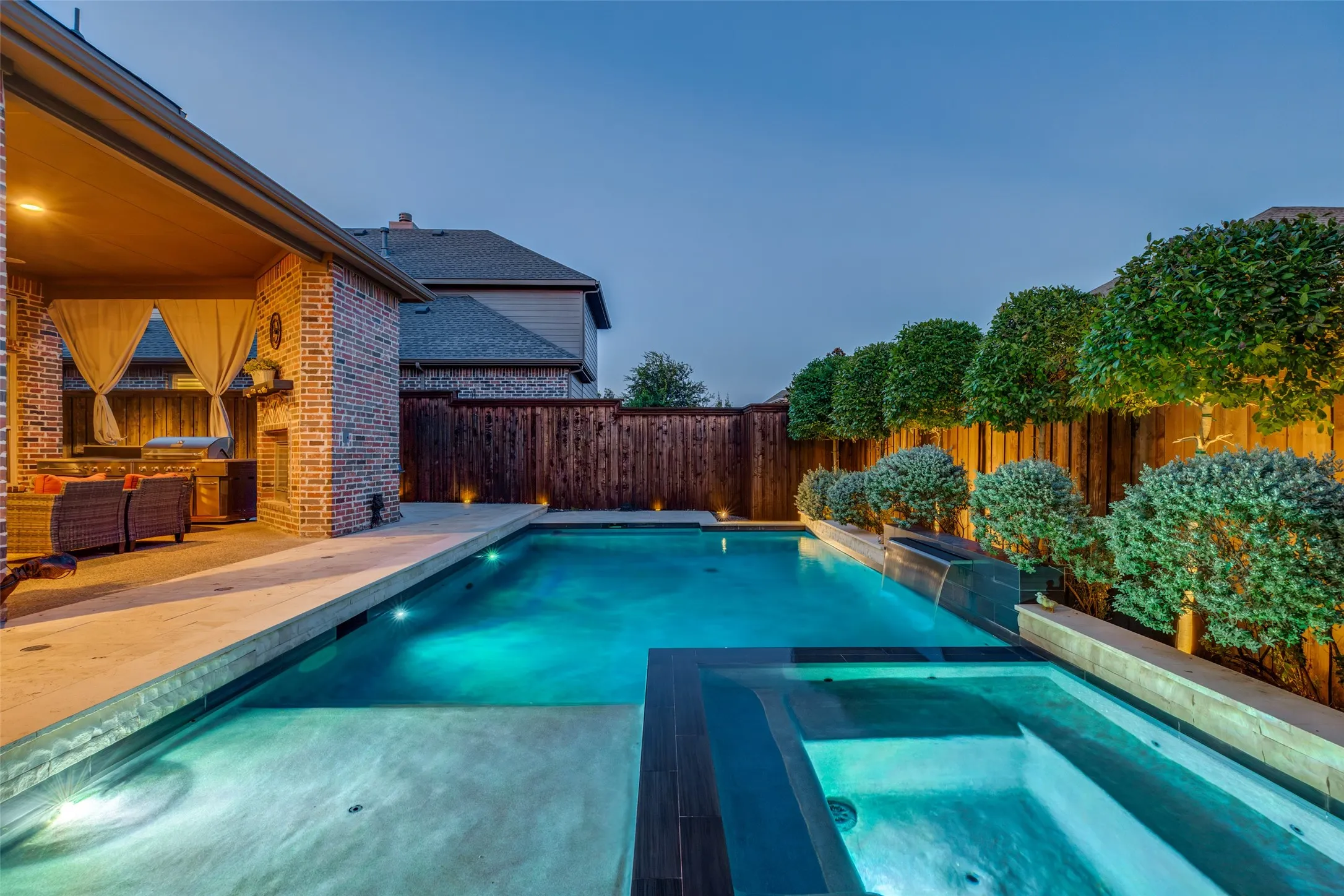 Outdoor living area featuring a covered patio with a built-in outdoor kitchen, a brick exterior, a swimming pool with integrated spa, and manicured landscaping