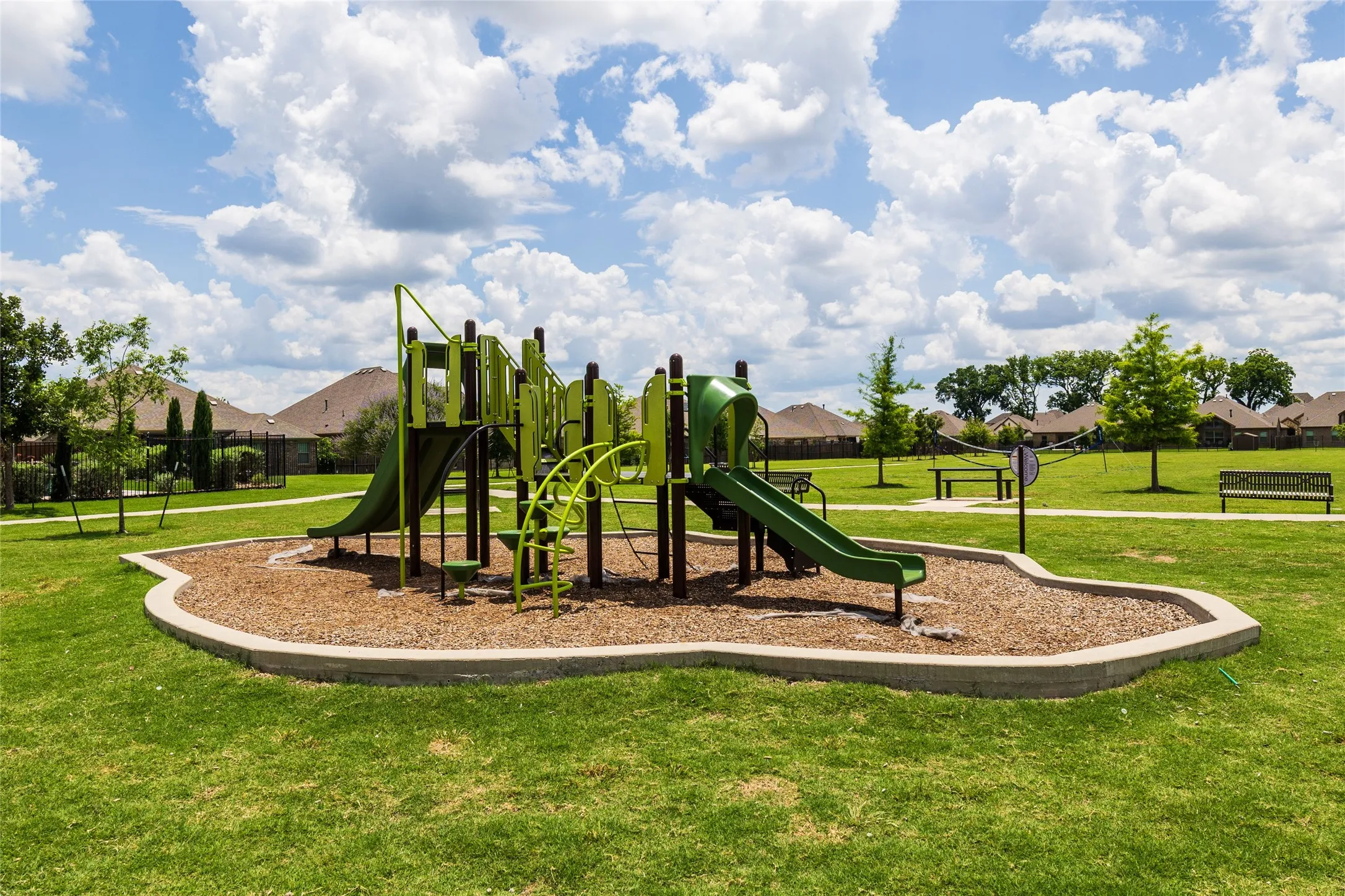 Community playground featuring a multi-level play structure with slides and climbing elements, surrounded by a wood chip surface and a concrete border