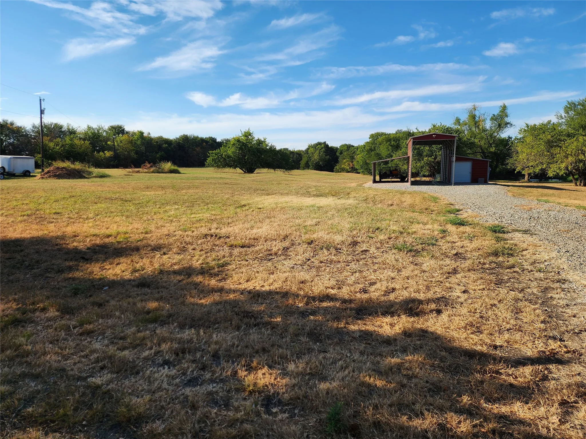 Expansive lot featuring a gravel driveway, open-air structure, and mature trees