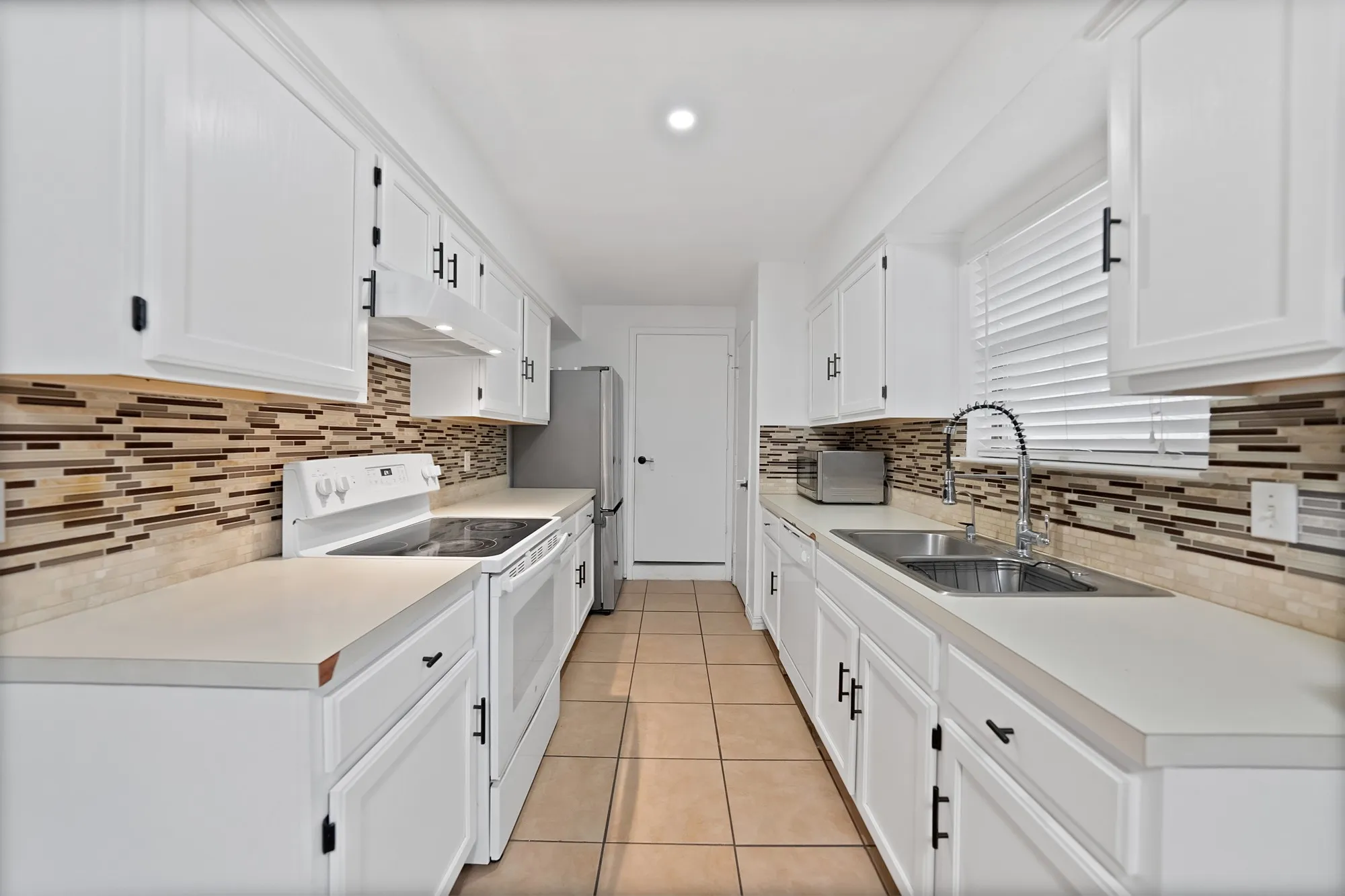 Galley kitchen featuring white cabinetry, light-toned countertops, a mosaic tile backsplash, stainless steel sink, and ceramic tile flooring