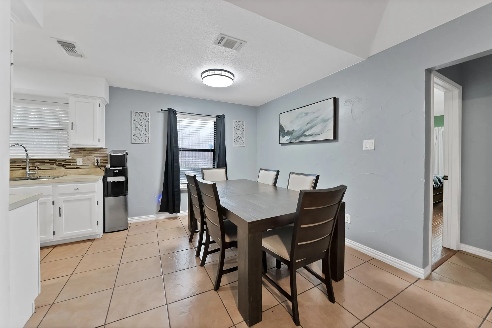 Open-concept kitchen and dining area featuring tile flooring, white cabinetry, a neutral-toned backsplash, and a large window