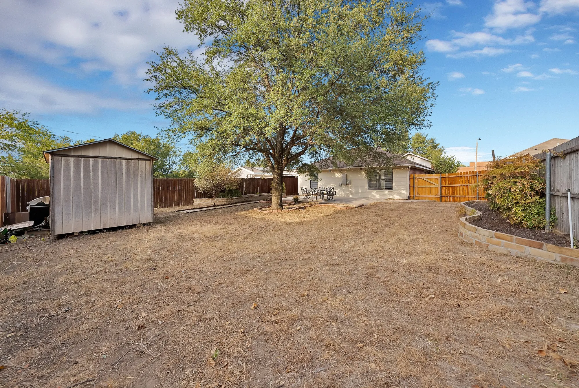 Expansive backyard featuring a mature tree, wood fencing, a storage shed, and a stone-edged garden bed