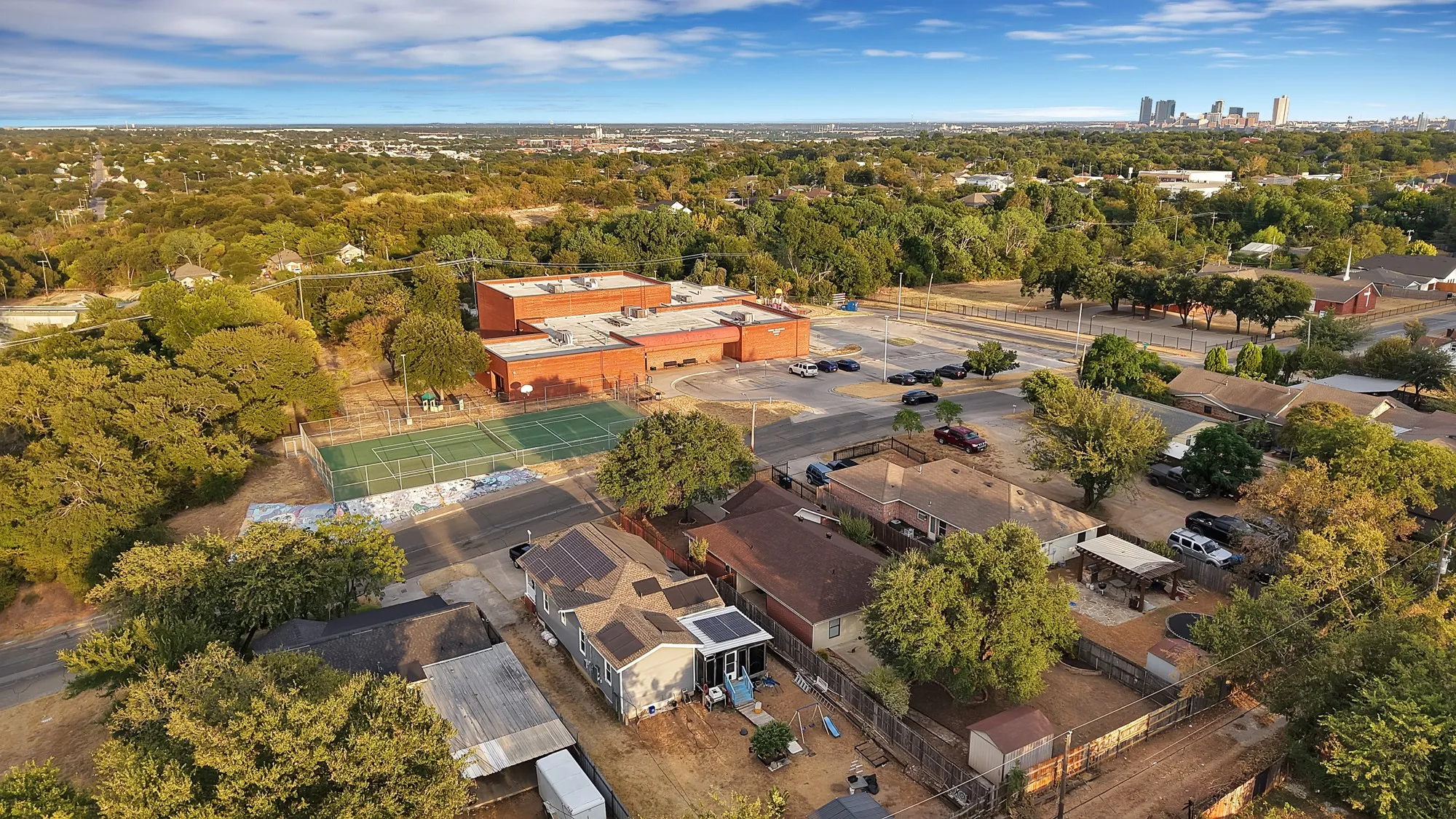 Aerial perspective showcasing surrounding residential properties with mature trees, a nearby brick building, and recreational courts
