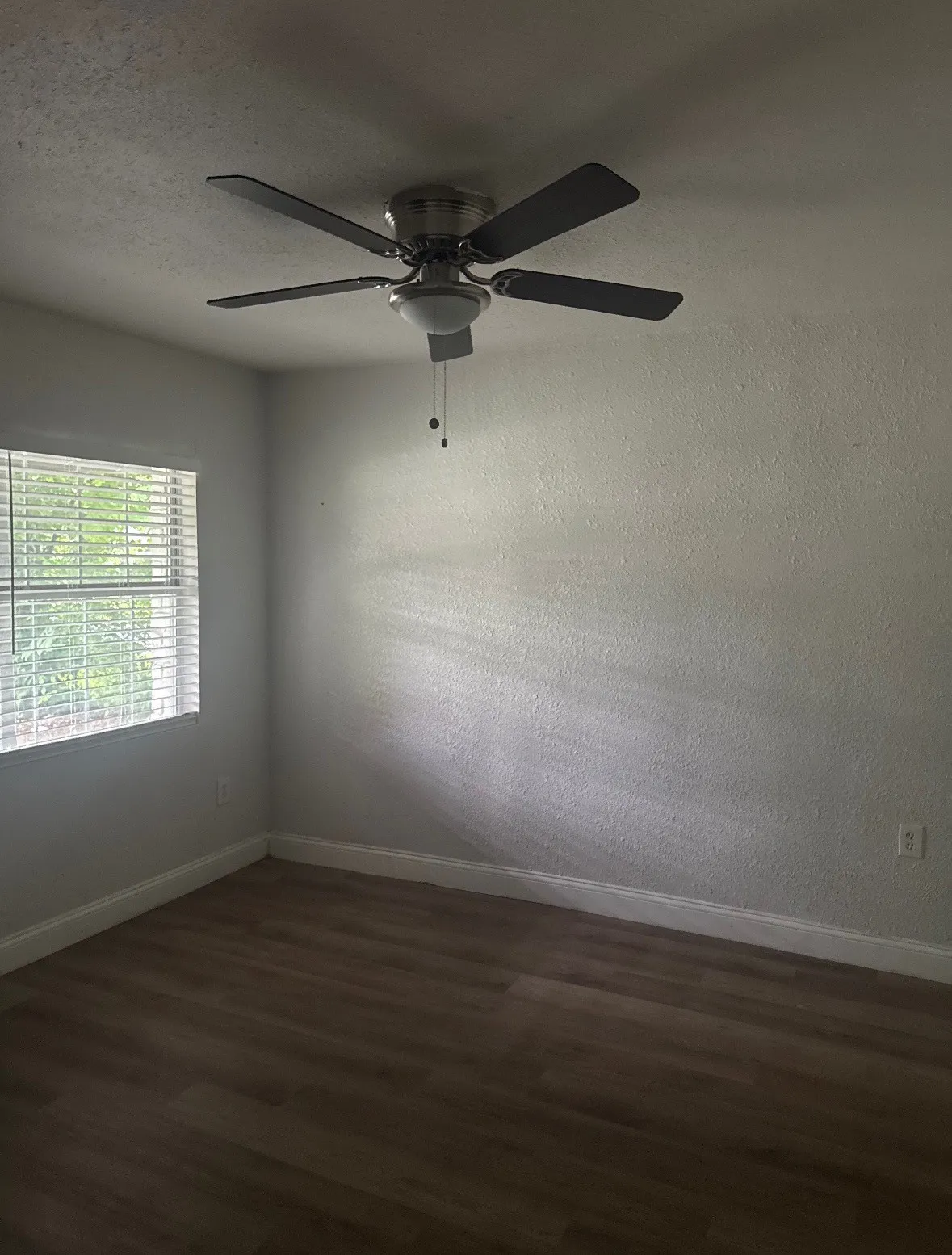 Wood-finish flooring, neutral wall tones, window with horizontal blinds, and a ceiling fan with integrated lighting