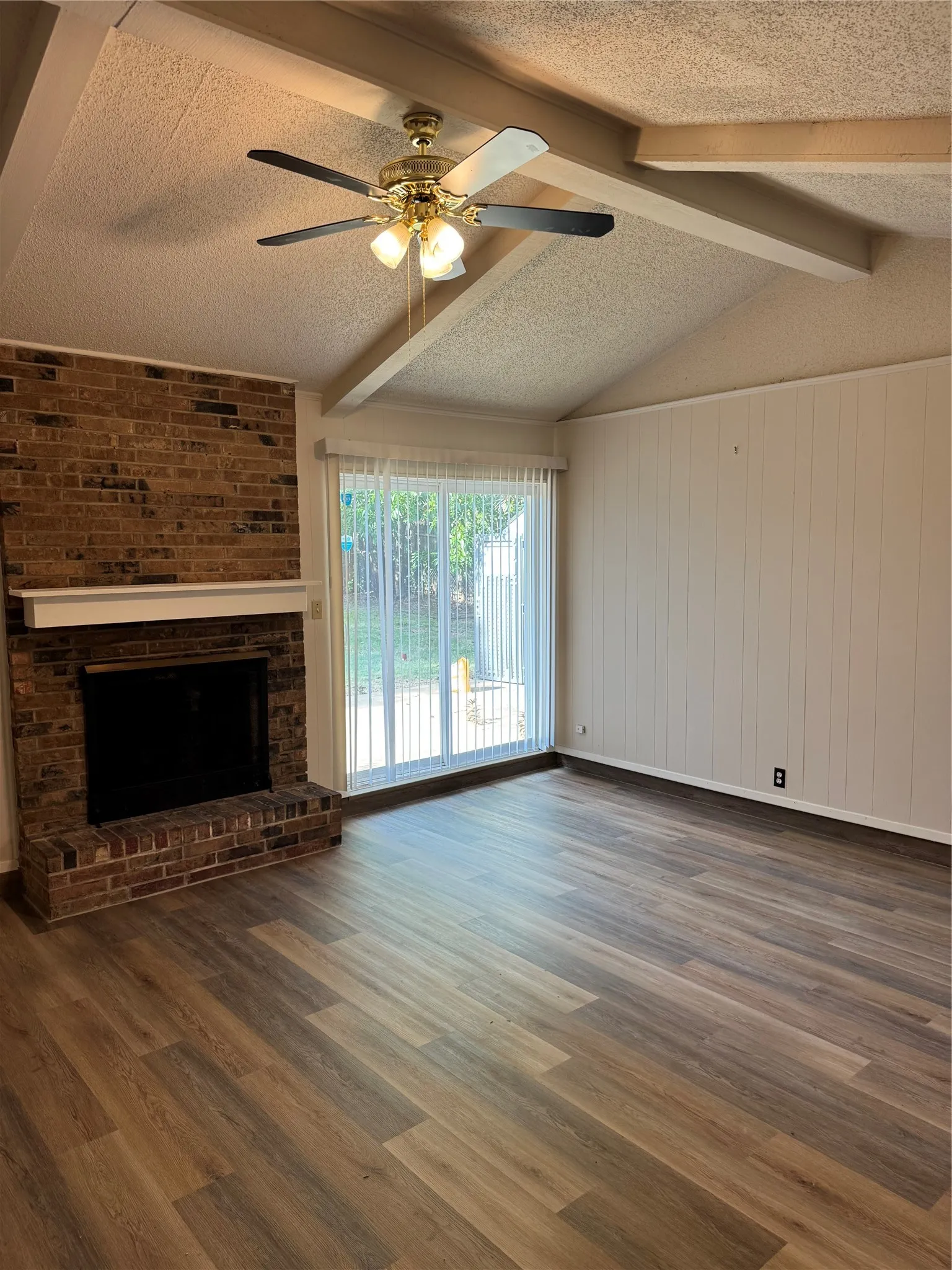 Vaulted ceiling with exposed beams, brick-faced fireplace with hearth, wood-finish flooring, and sliding glass door