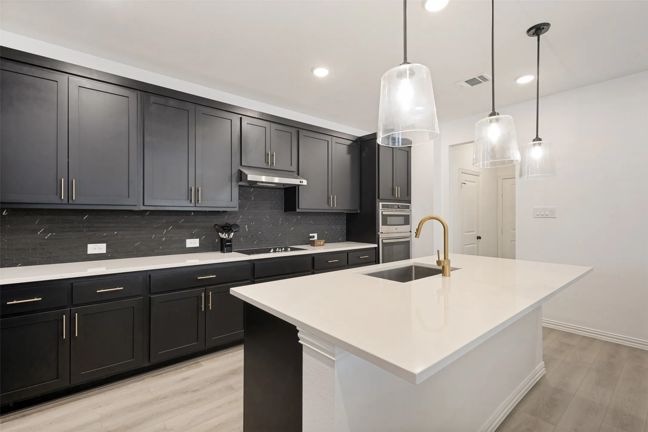 Kitchen featuring dark cabinetry, white countertops, built-in stainless steel appliances, a center island with an undermount sink, and three pendant lights