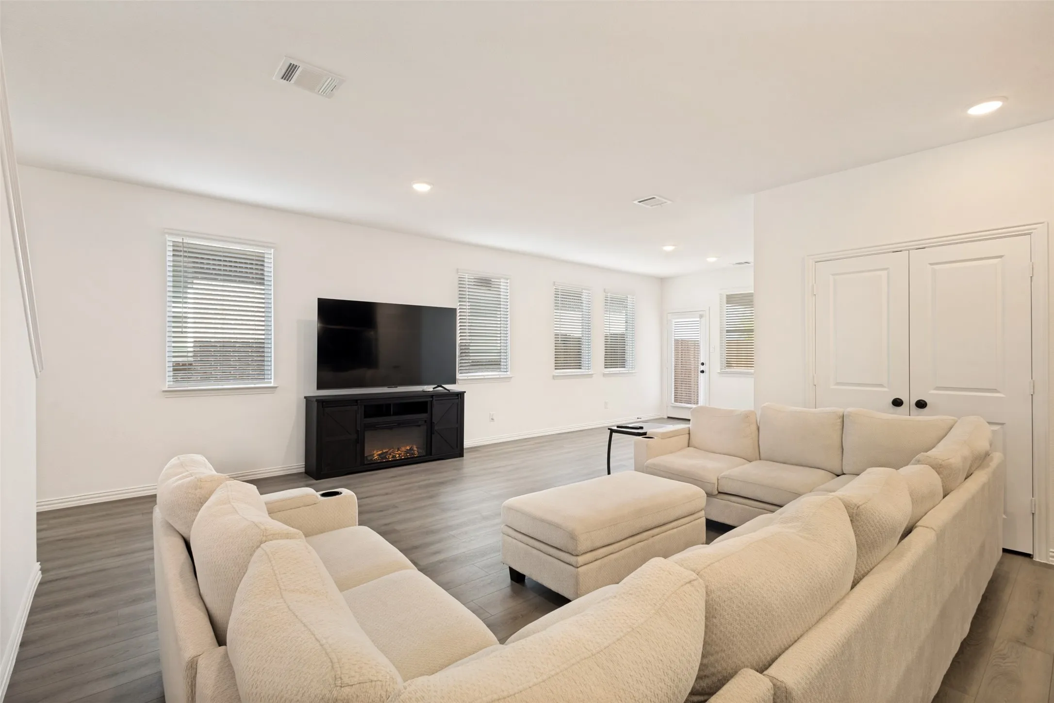 Spacious living area featuring wood-finish flooring, recessed lighting, multiple windows with blinds, and white double doors