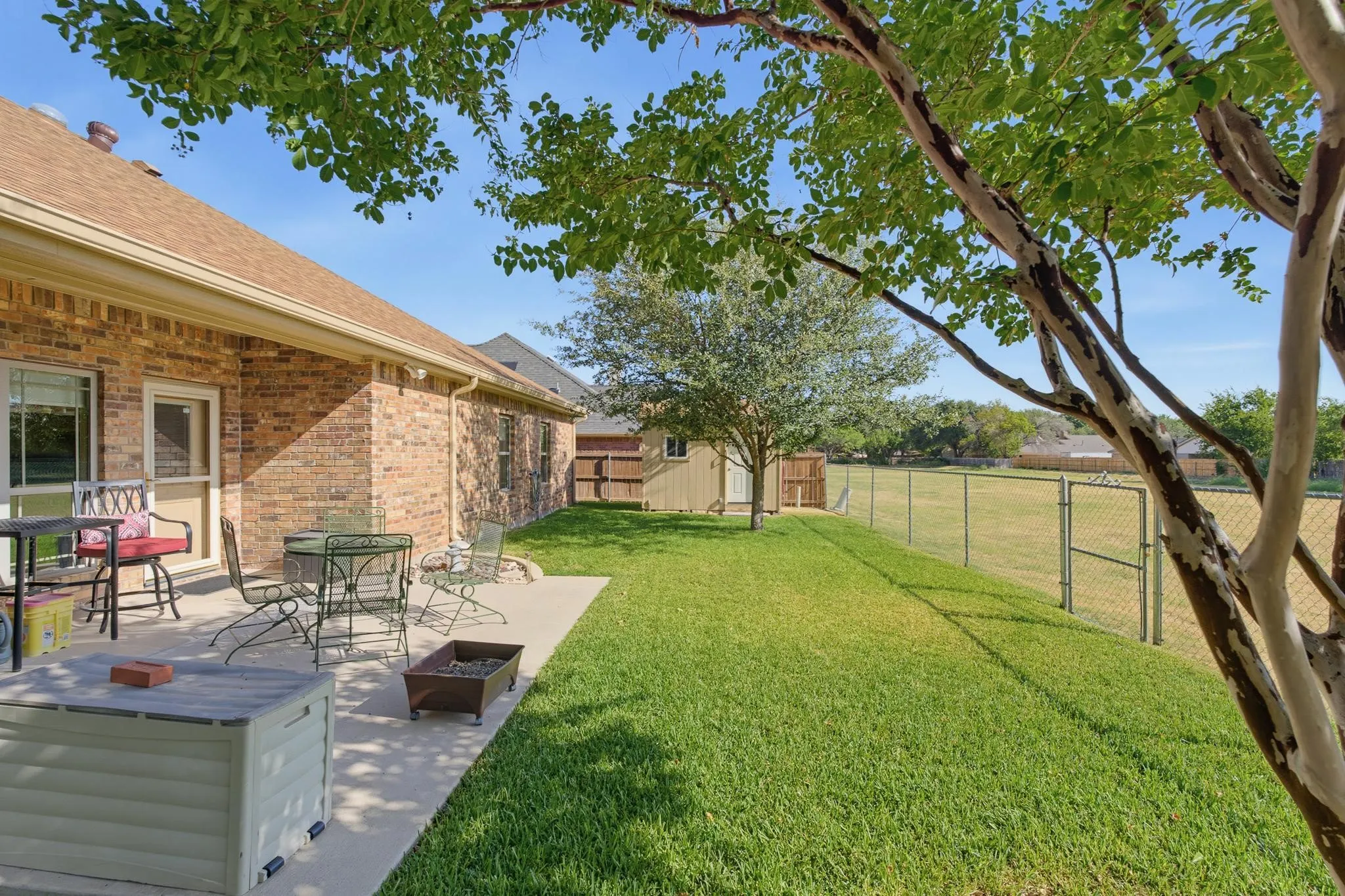 Brick exterior, concrete patio, expansive lawn, chain-link fencing, and mature trees