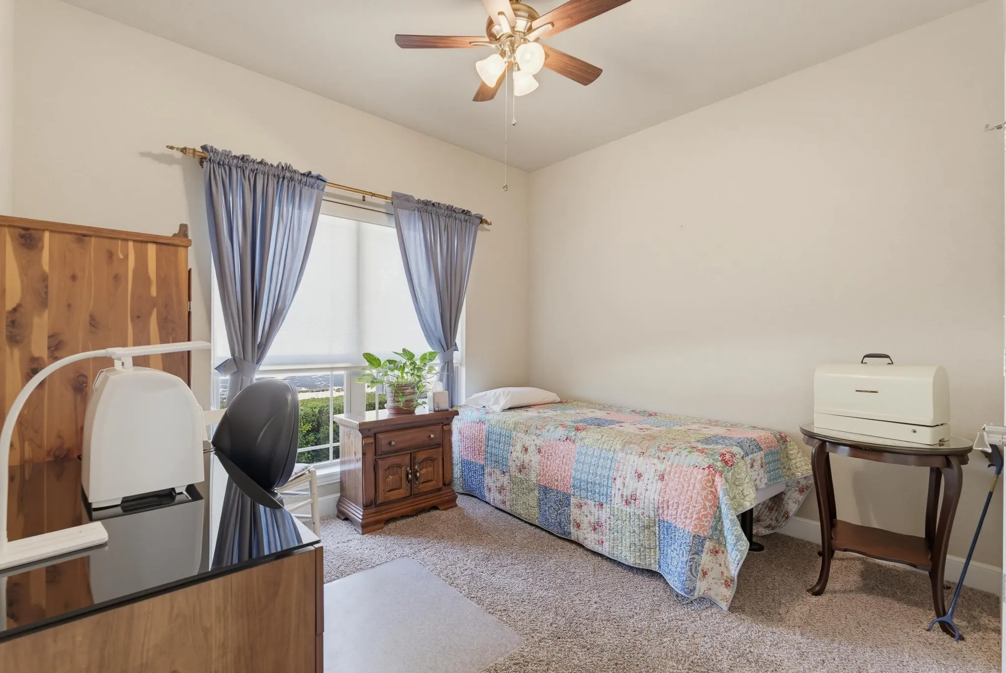 Carpeted flooring, window with blinds and drapes, wood-finish desk, wood-finish armoire, and ceiling fan with light fixture