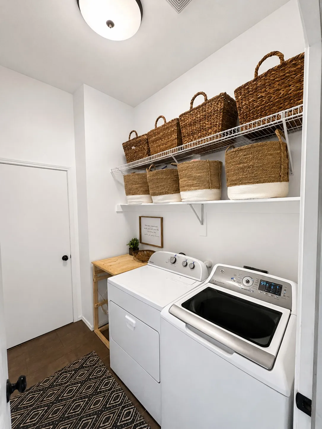 Dedicated laundry area featuring white walls, durable flooring, a built-in wire shelving system, a ceiling-mounted light fixture, and a solid white door with black hardware