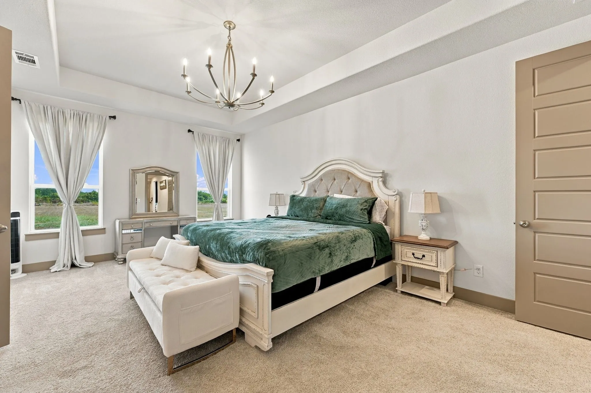 Recessed ceiling with an ornate chandelier, two large windows, light-colored carpeting, and a five-panel interior door