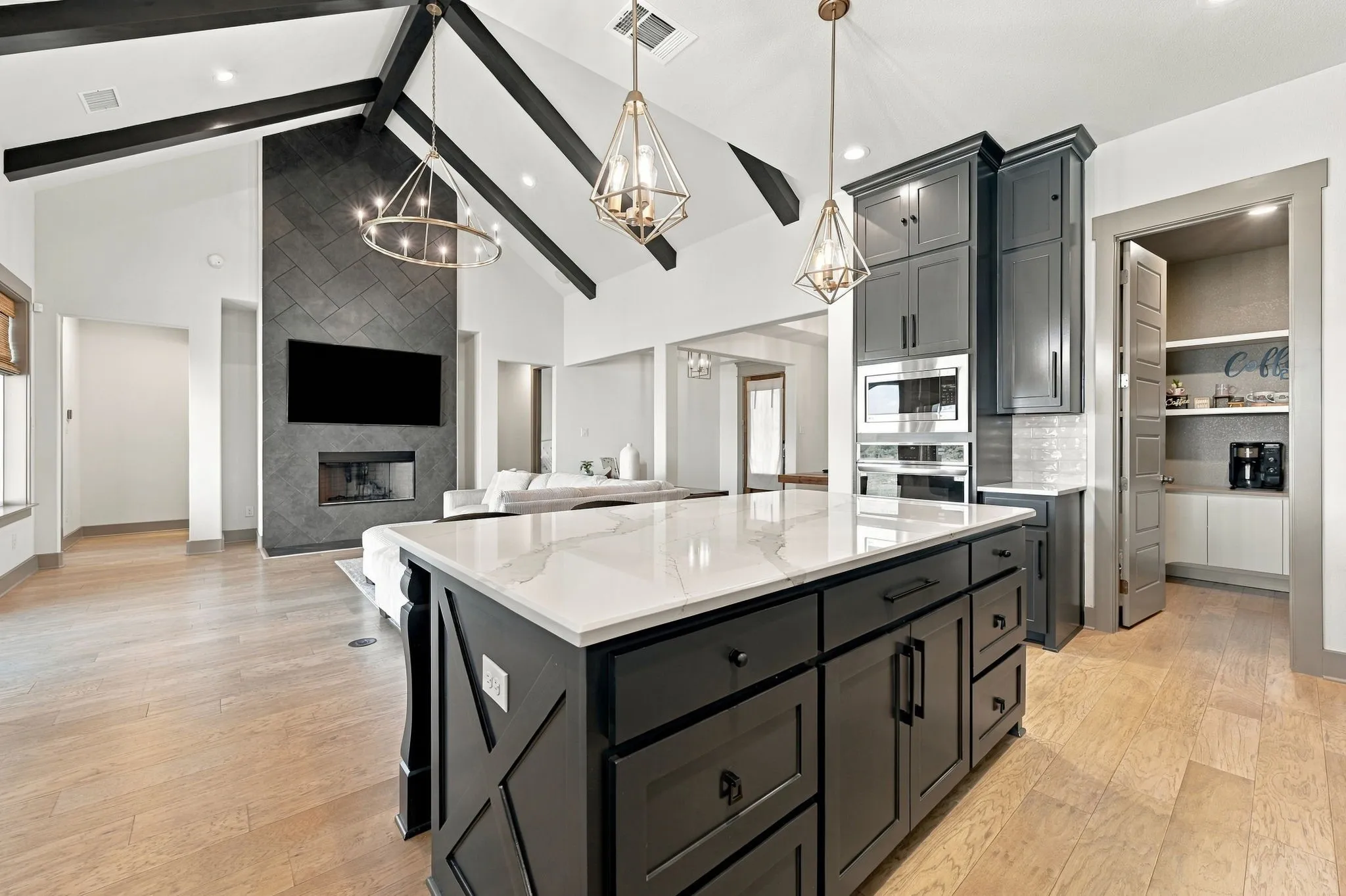 Vaulted ceilings with exposed beams, wood-finish flooring, a kitchen island with a light-toned countertop, built-in appliances, and a pantry with shelving