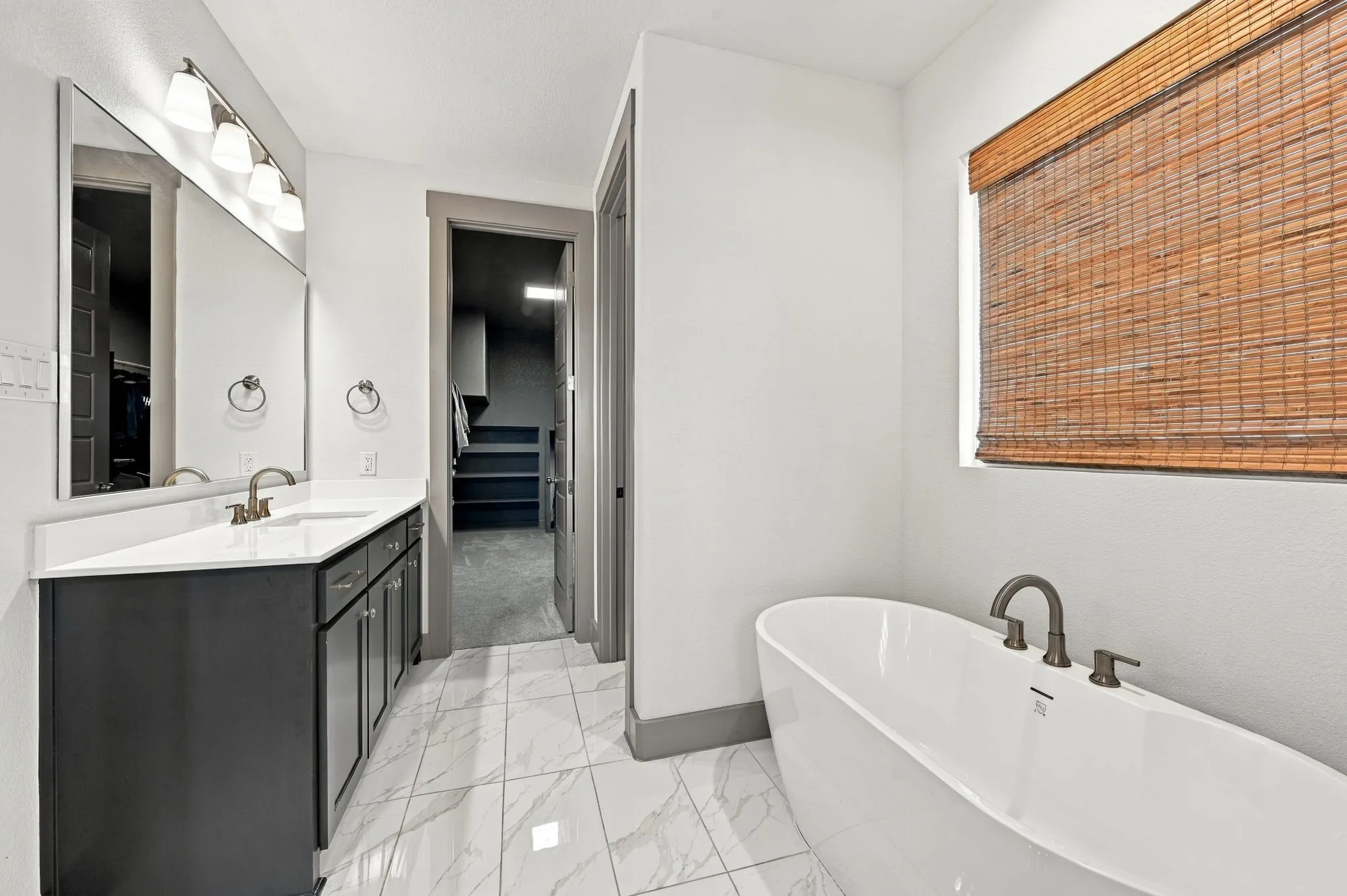 Freestanding soaking tub with dark-toned fixtures, wood-slat window treatment, white tile flooring, and a vanity with a white countertop and dark cabinetry