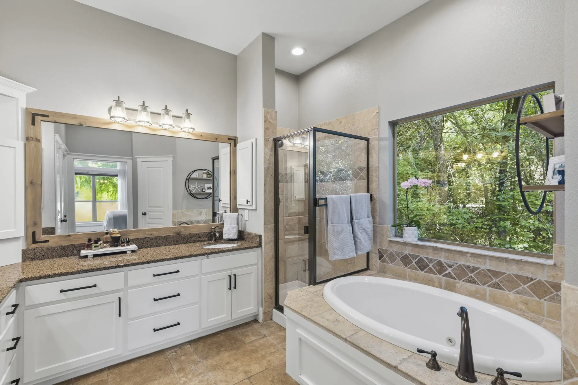 Dual vanity with white cabinetry and dark hardware, framed mirror with integrated lighting, tiled flooring, built-in soaking tub with dark fixtures, and a glass-enclosed shower