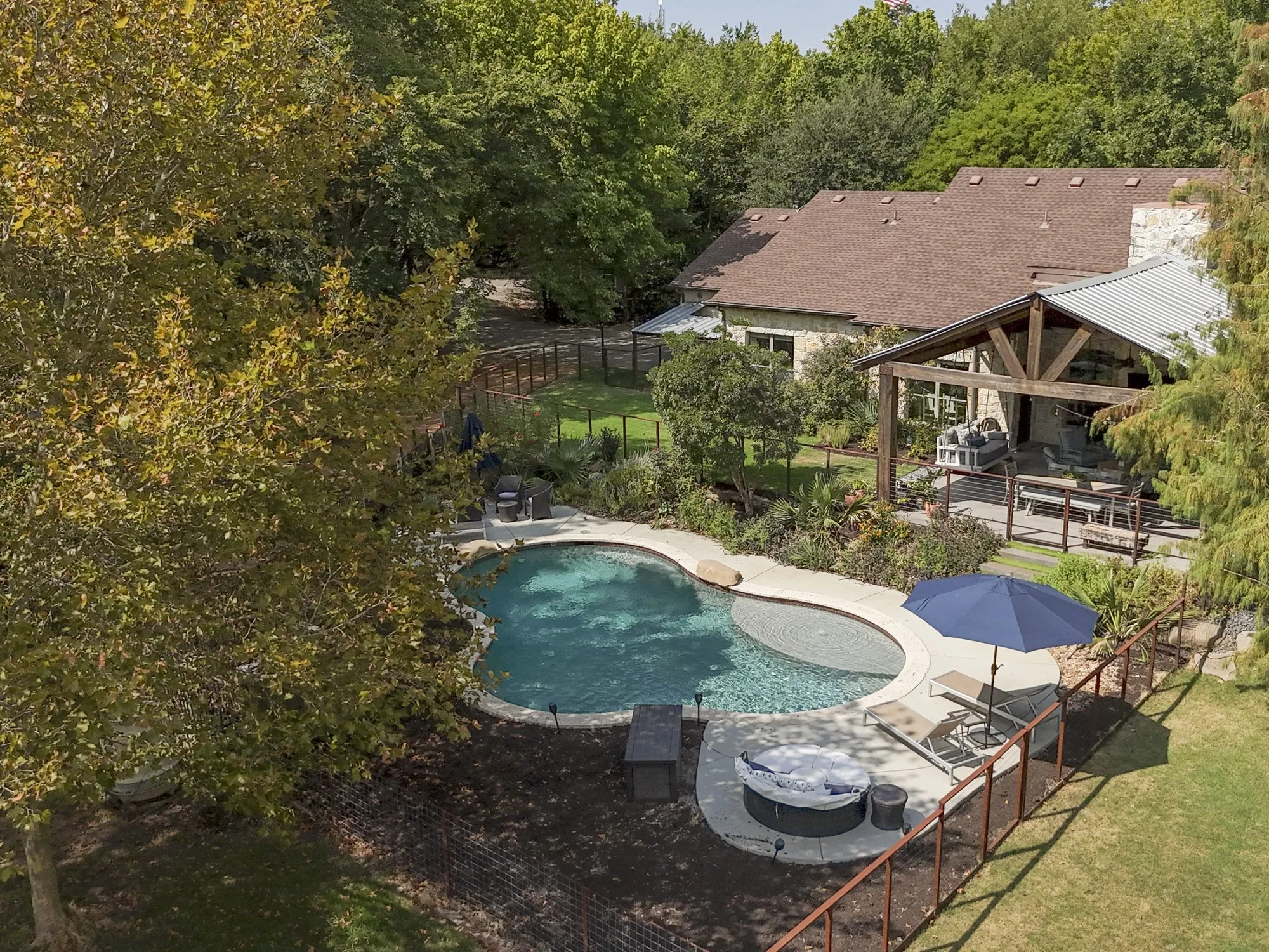 Custom-shaped swimming pool with an integrated spa, expansive covered patio with exposed beam trusses, and a stone-clad exterior