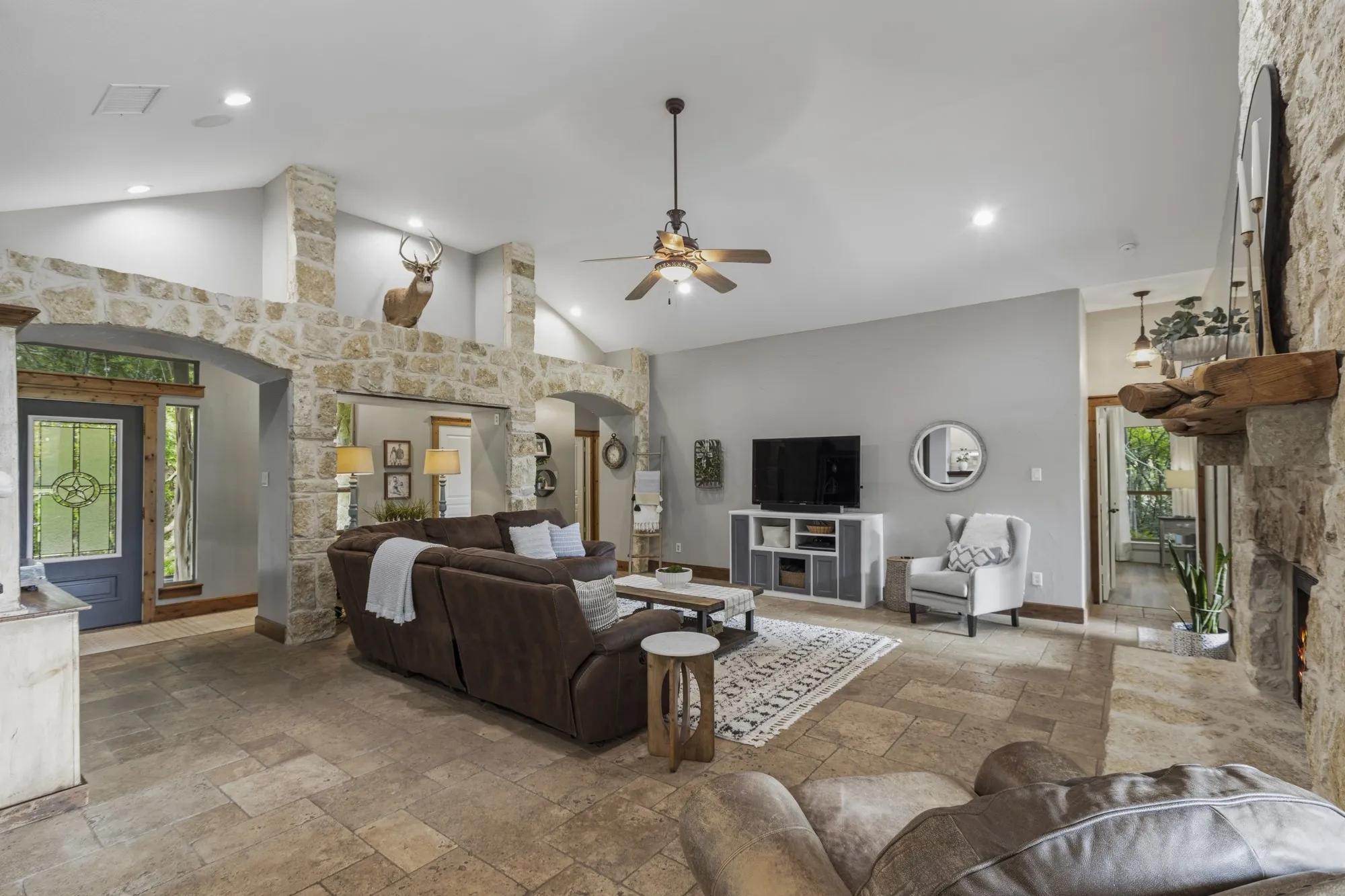 Vaulted ceiling with recessed lighting and ceiling fan, stone-clad fireplace with wood mantle, tile flooring, stone archways, and a built-in media cabinet