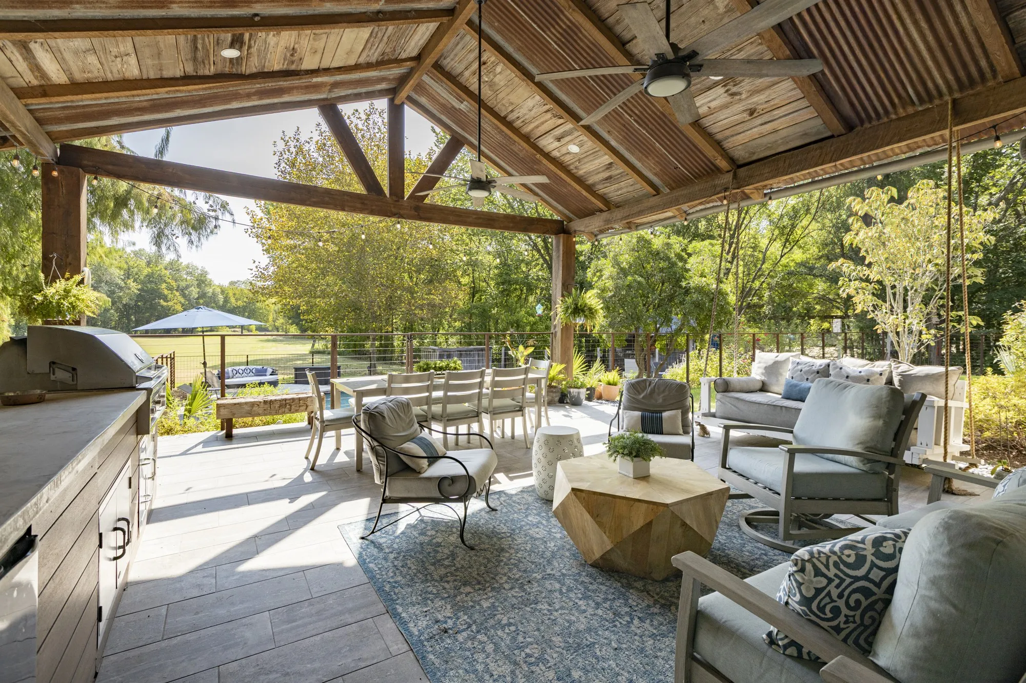 Spacious covered patio with wood-beamed ceiling and corrugated metal accents, featuring an outdoor kitchen, ceiling fans, and stone-finish flooring