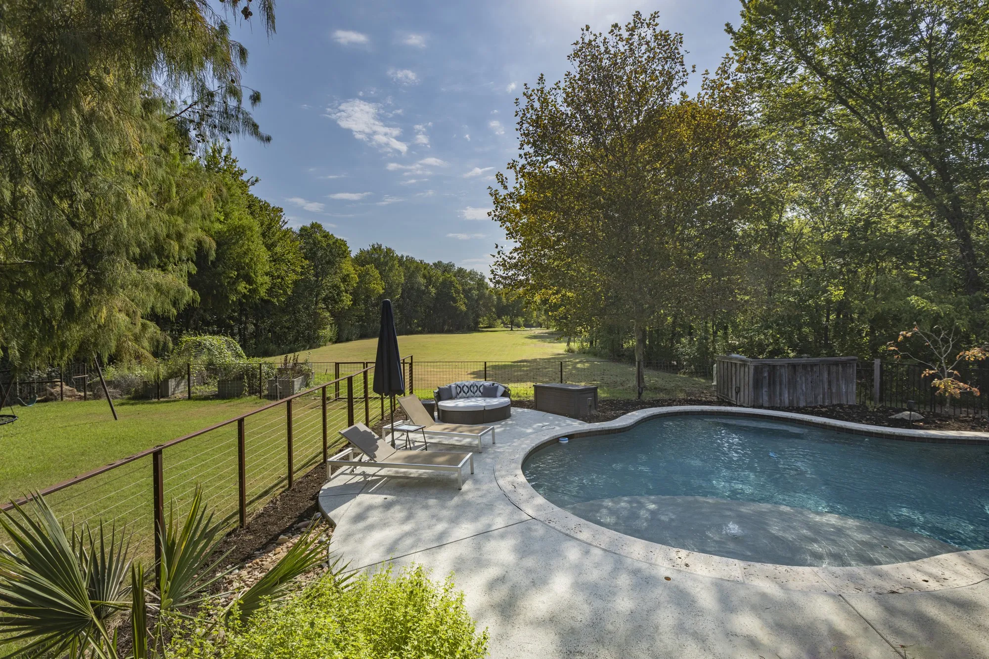 Curved swimming pool with a concrete surround, wood-paneled privacy screen, and a wire-cable fence