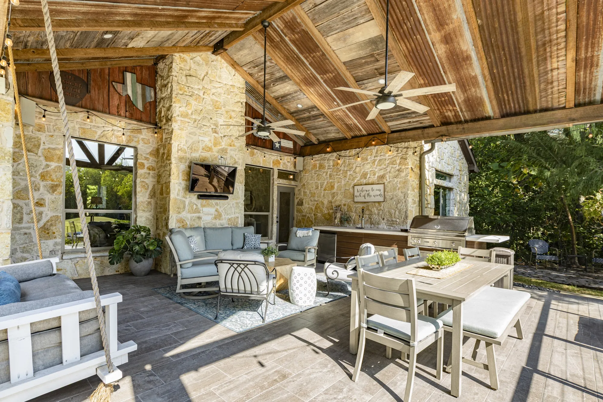 Expansive covered patio featuring a stone exterior, wood-paneled ceiling, ceiling fans, and an outdoor kitchen with an integrated grill