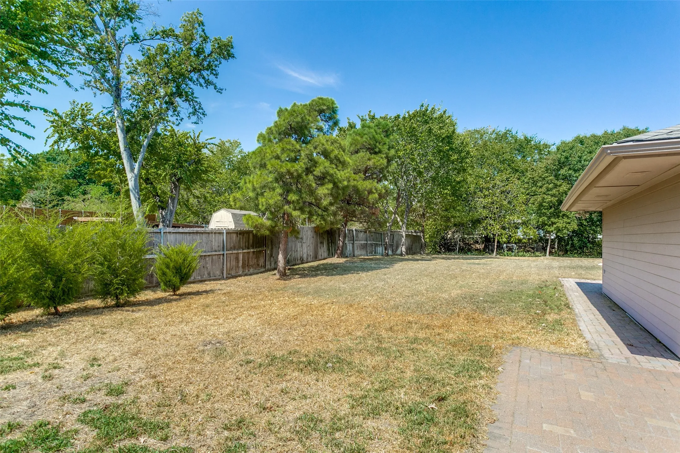 Expansive backyard featuring a privacy fence, mature trees, and a paved patio area