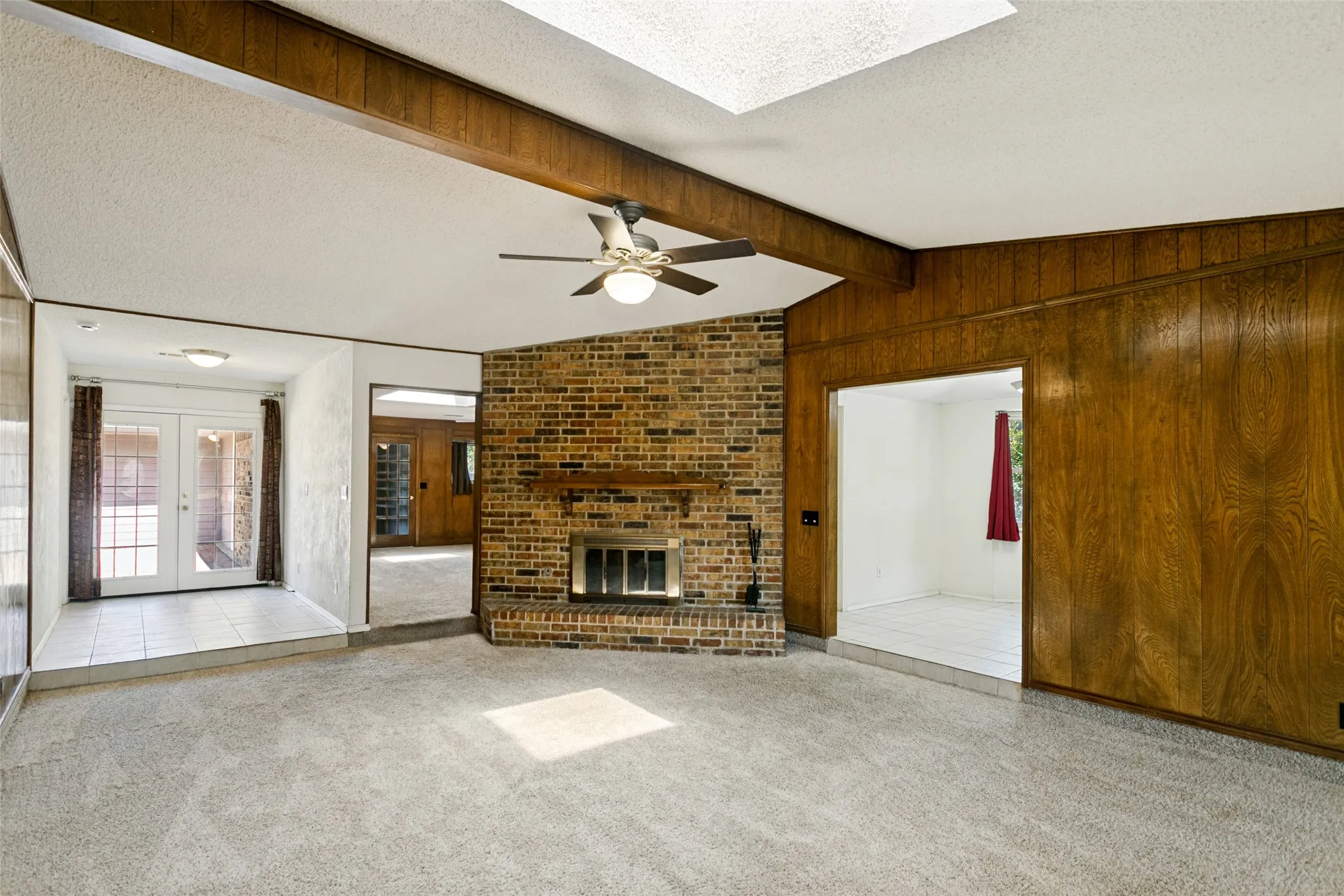 Brick fireplace with wood mantel, wood paneling, ceiling fan with integrated lighting, and multiple skylights