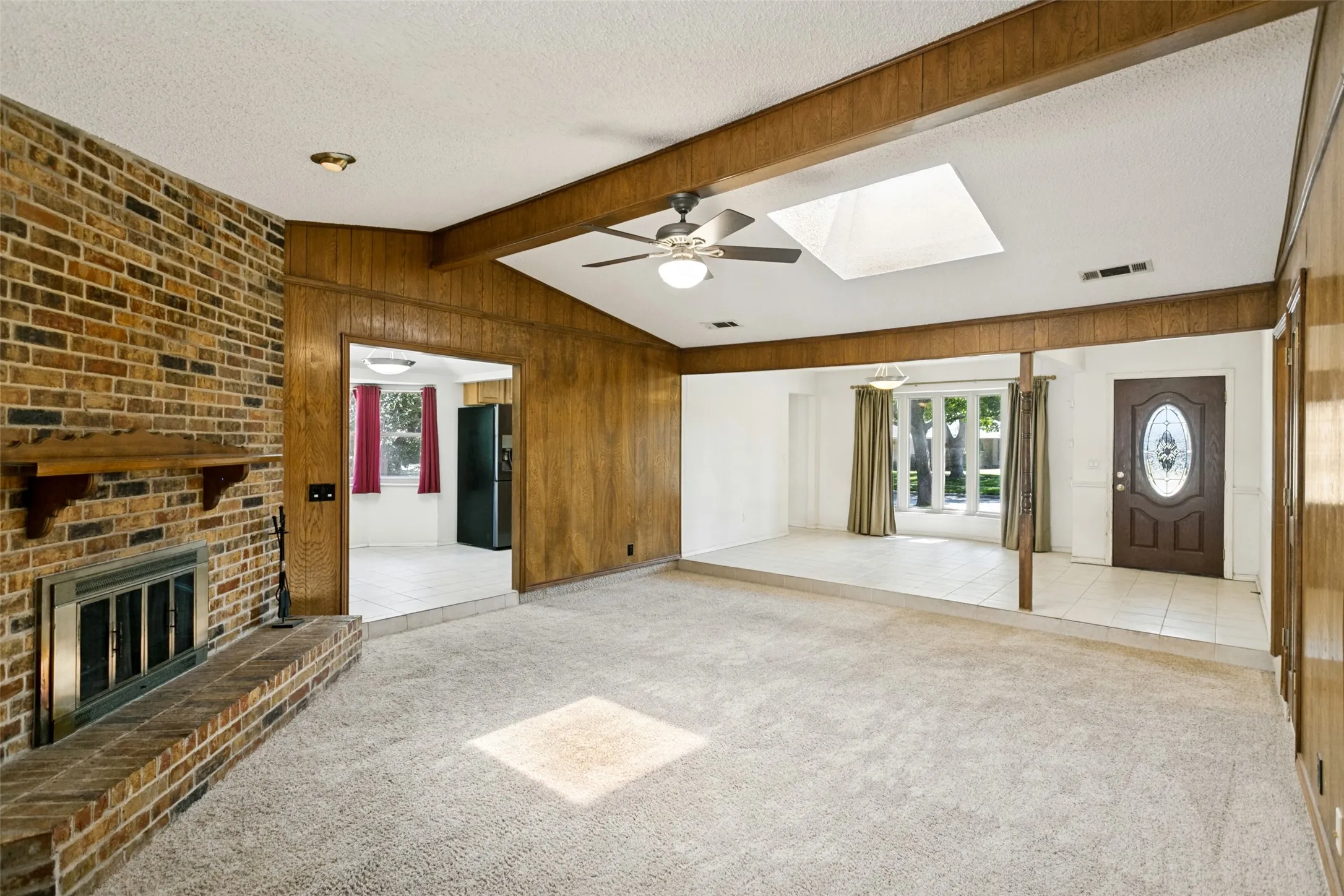 Vaulted ceiling with a skylight and ceiling fan, exposed brick fireplace with a hearth and wood mantel, wood paneling, and a decorative front entry door