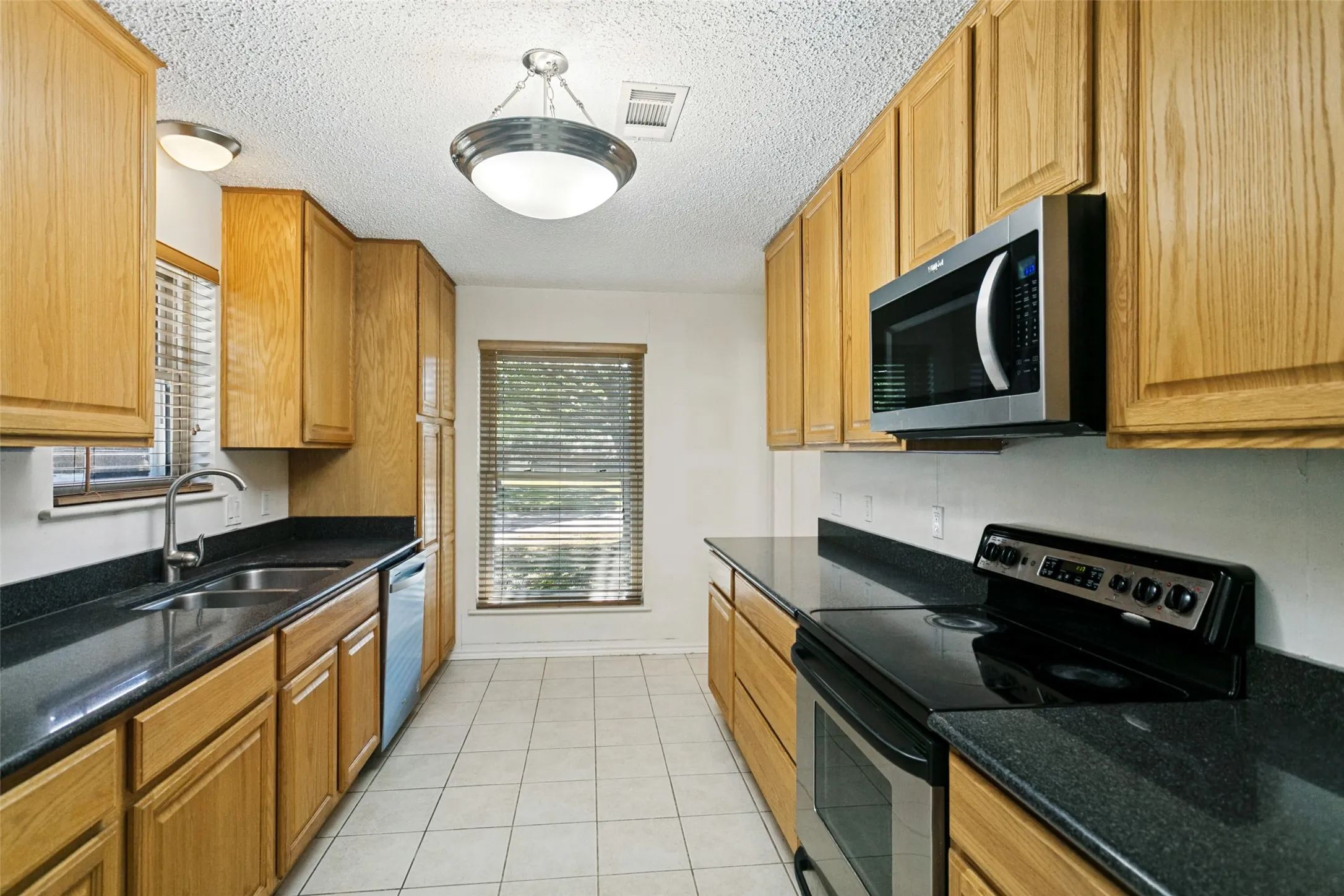 Galley kitchen featuring wood-finish cabinetry, dark countertops, and stainless steel appliances including a microwave and range