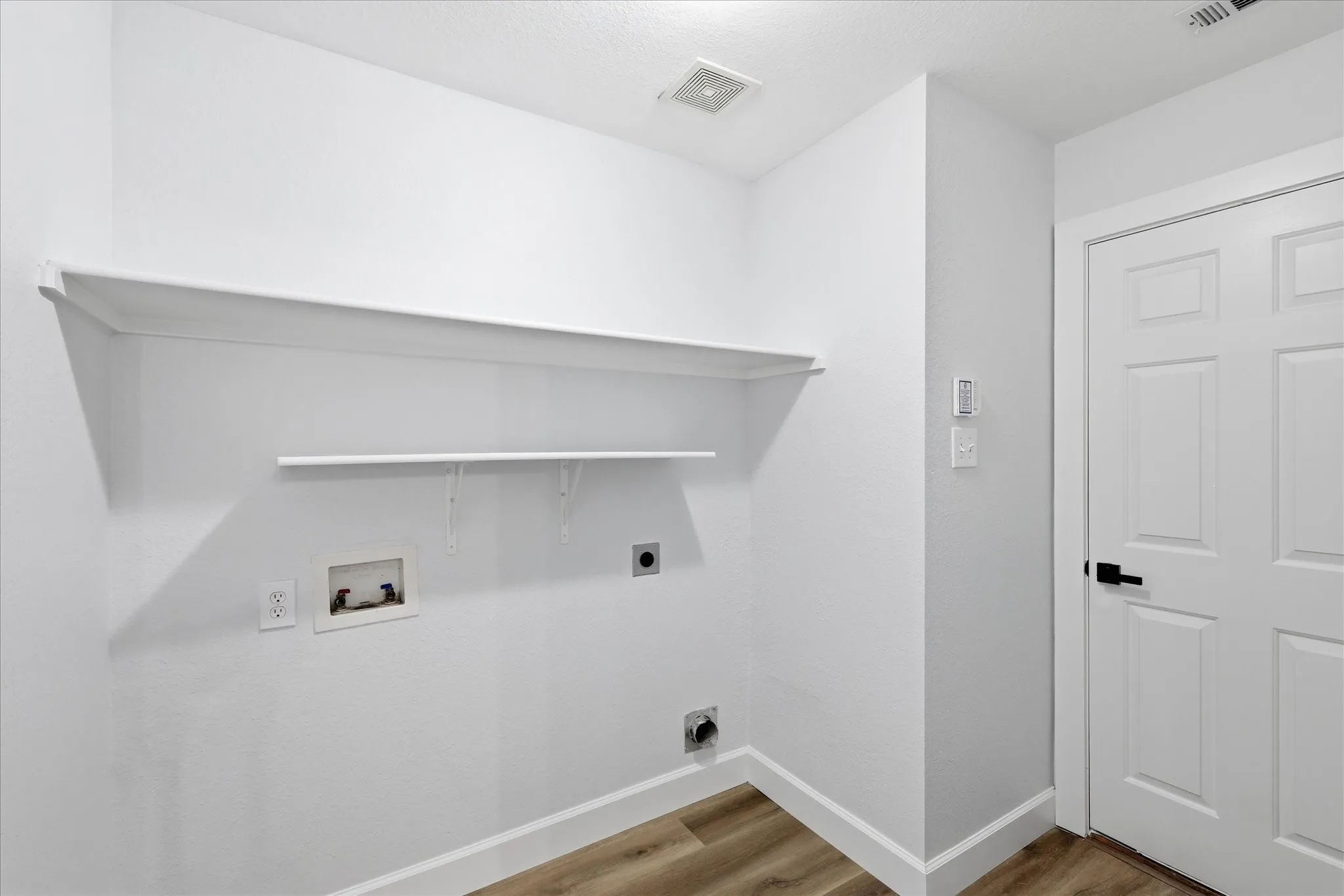 Laundry room featuring two wall-mounted shelves, wood-finish flooring, and a white panel door with black hardware
