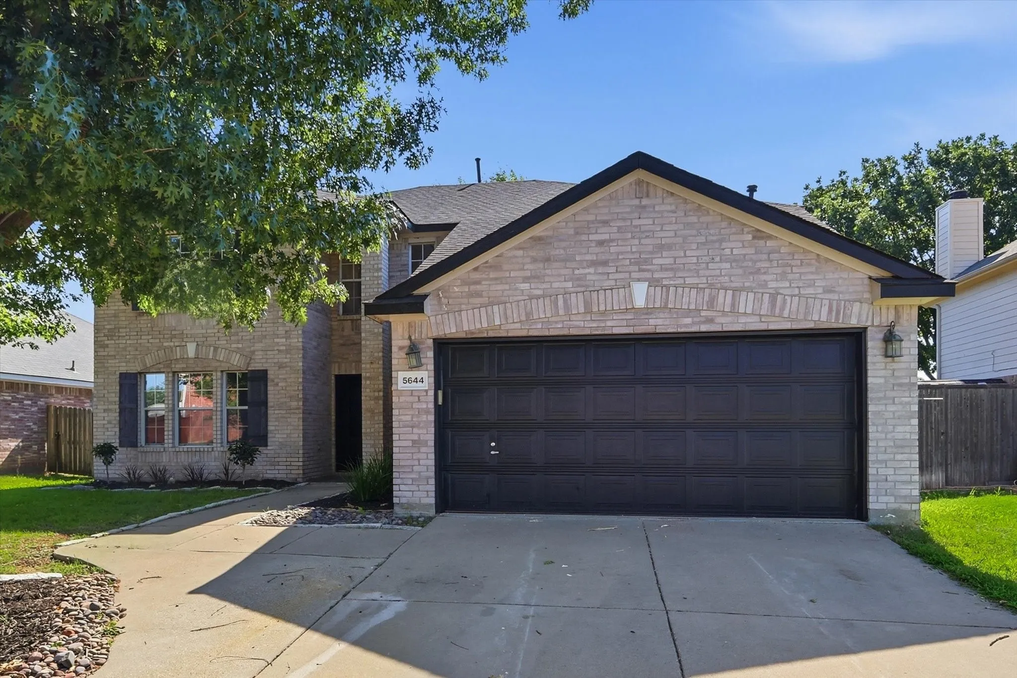 Contemporary brick home featuring a spacious driveway with concrete finish, black aluminum garage door, gabled roofline with architectural detailing, and mature landscaping with established trees framing the front elevation