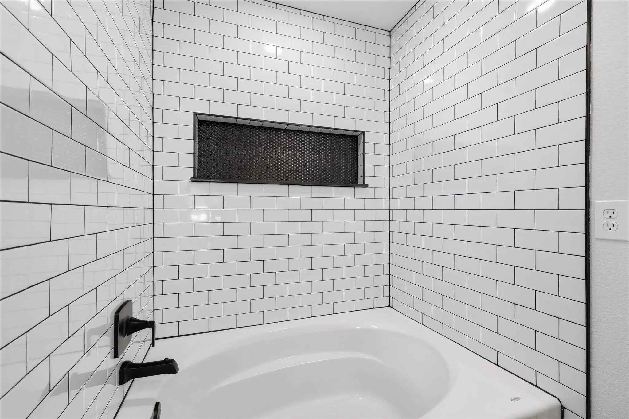 Bathtub with subway tile surround, built-in niche with dark mesh accent, and matte black fixtures