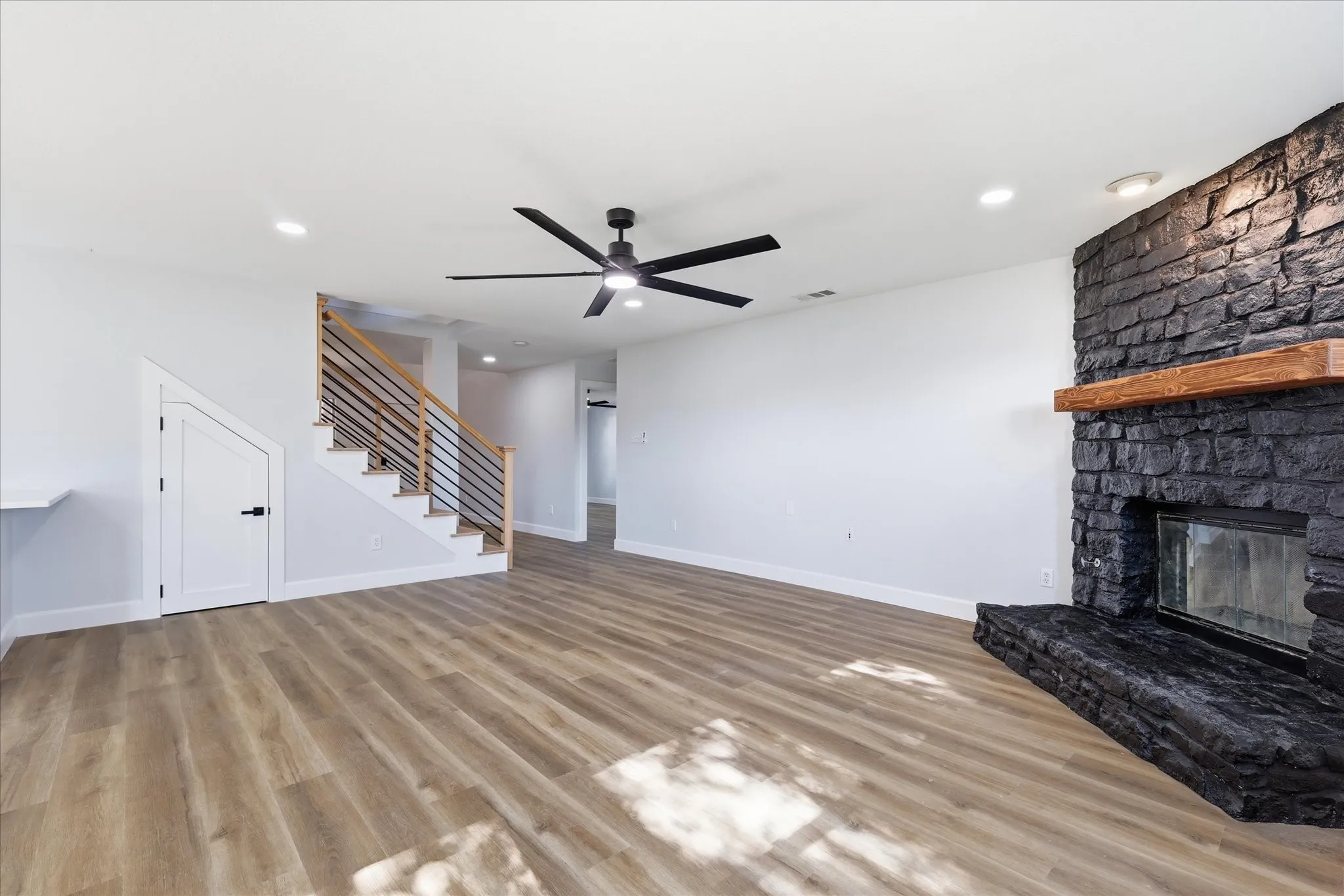 Living area featuring wood-finish flooring, a stone-clad fireplace with a wood mantle, a modern ceiling fan, recessed lighting, and a staircase with metal railings