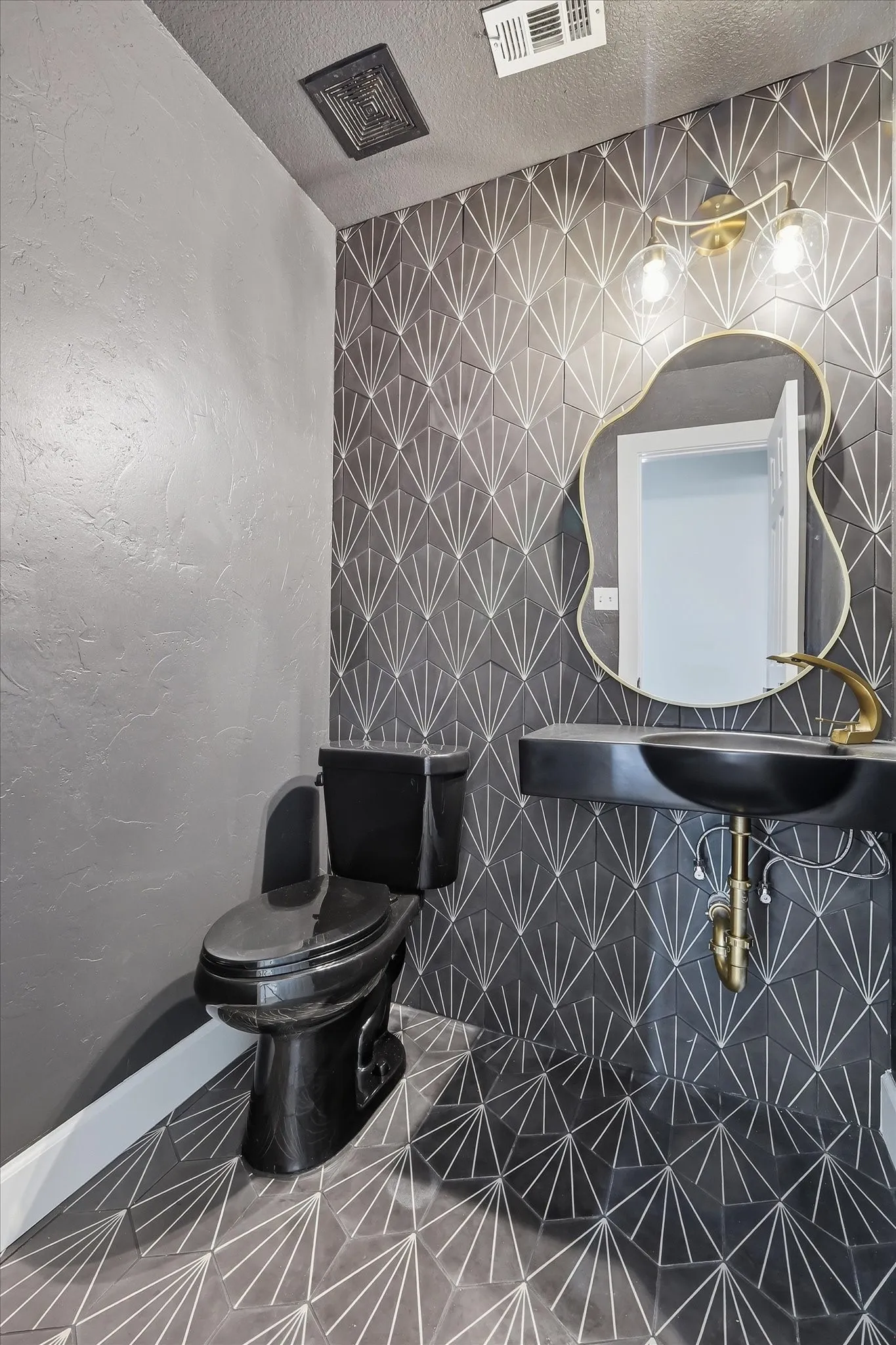 Geometric tile accent wall and flooring, black pedestal sink, black toilet, gold-finish faucet, and brass wall sconce