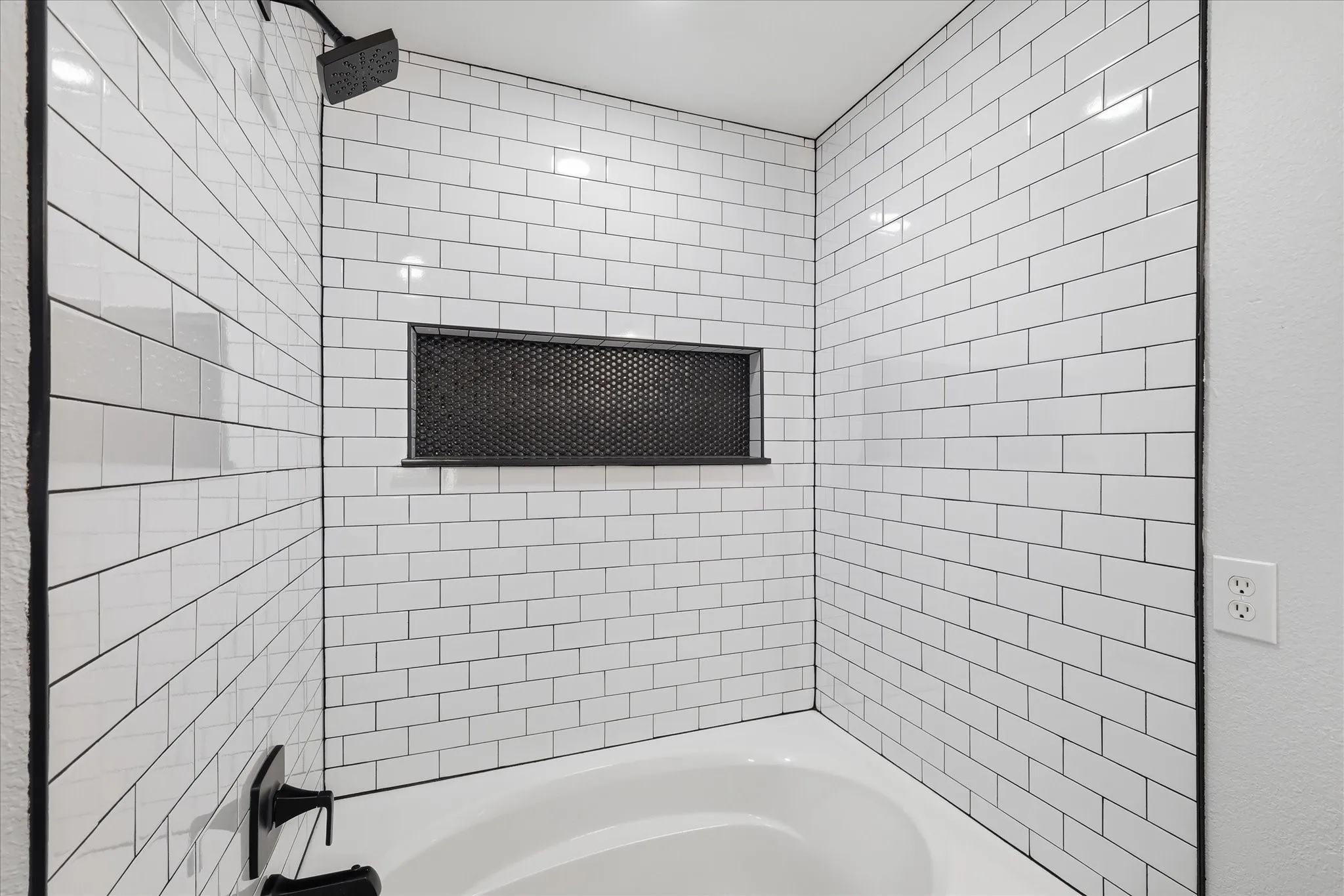Bathtub with subway tile surround, recessed niche, matte black shower fixtures, and dark grout lines